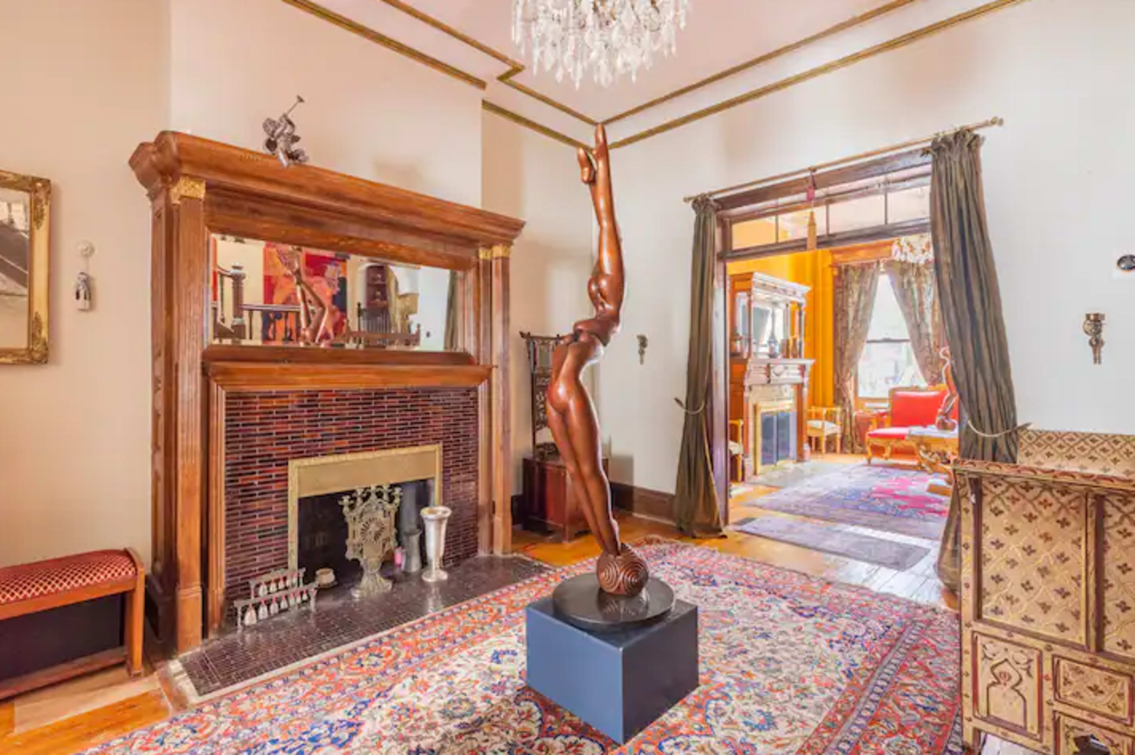 A spacious interior featuring a tall bronze sculpture on a pedestal, a wooden fireplace with a mirrored mantel, and an ornate rug, with a glimpse of an adjacent room through an open doorway.