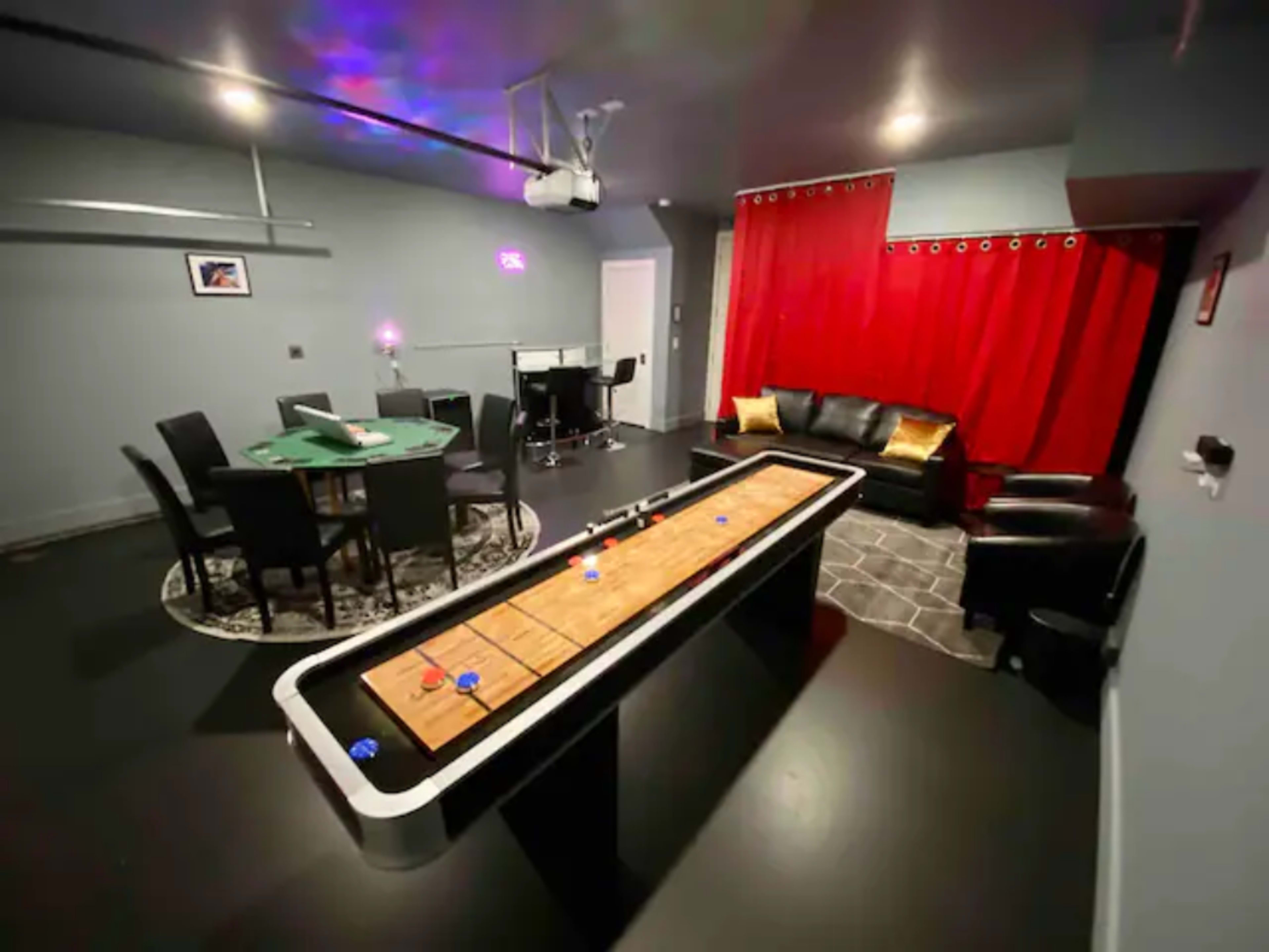 The image shows a game room featuring a shuffleboard table, a dining area with a circular table and chairs, a sitting area with a black sofa and chairs, and red curtains on one wall.