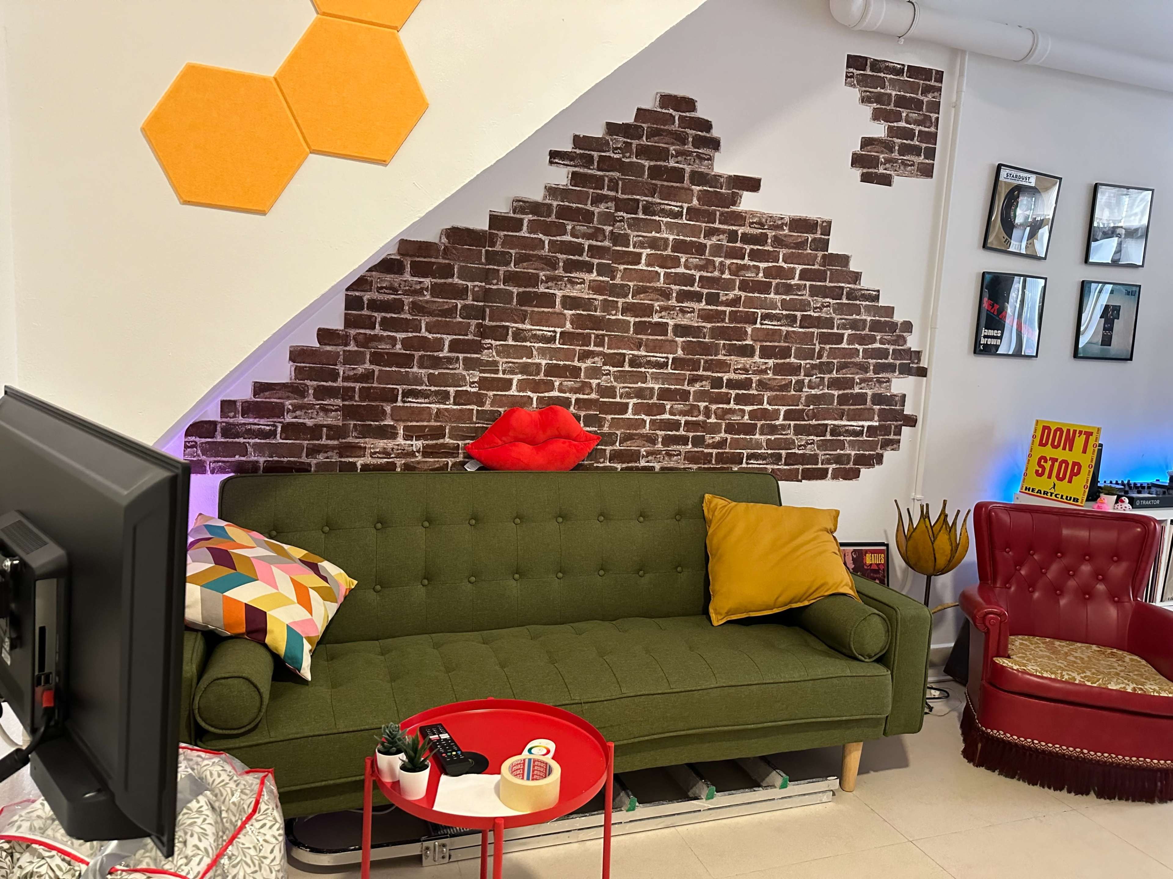 A green sofa with decorative cushions sits in front of a brick-patterned wall, accompanied by a red accent table and a vintage red chair.