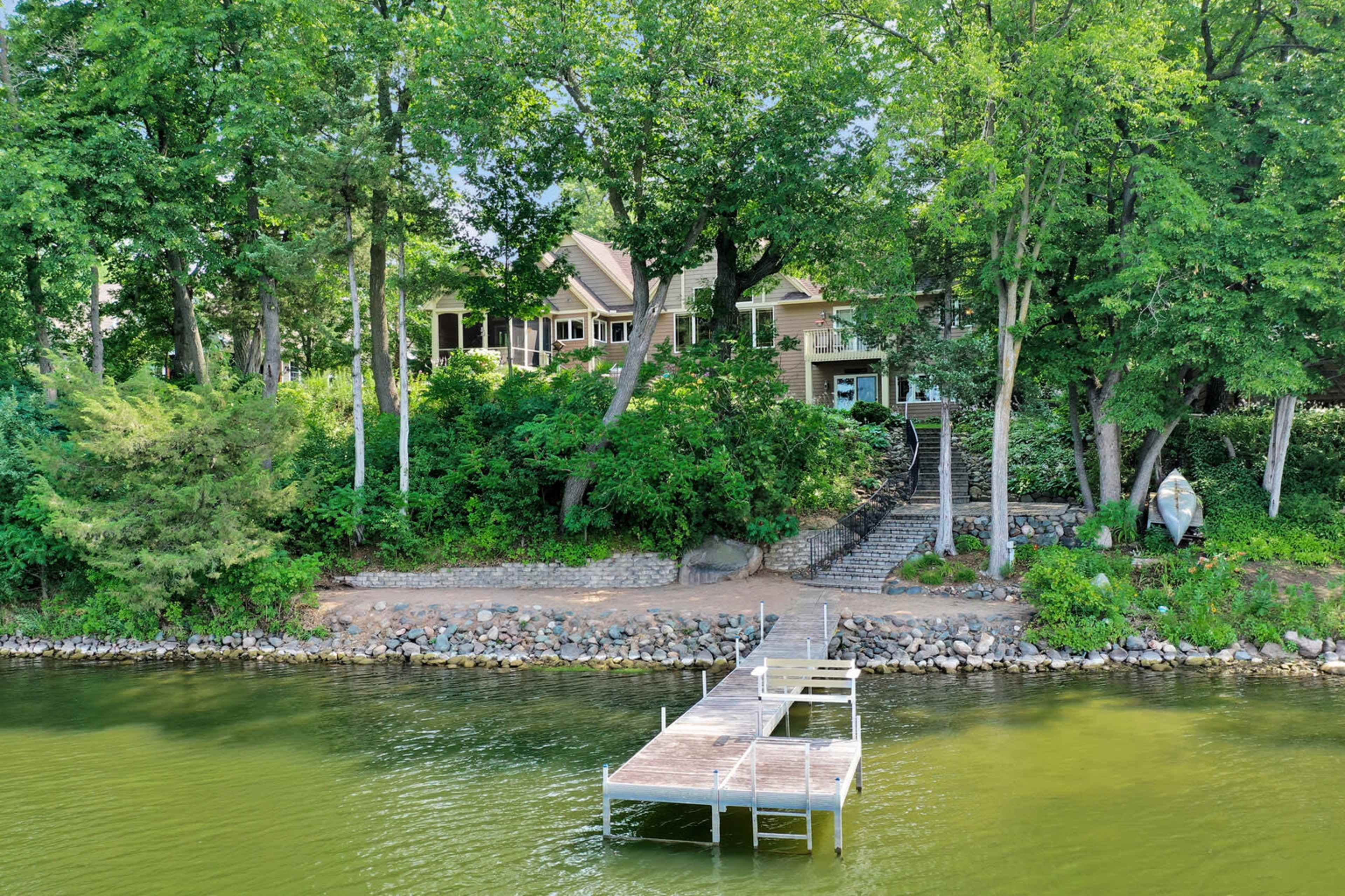 A wooden dock extends into a calm body of water, with a house and lush greenery visible along the shoreline.
