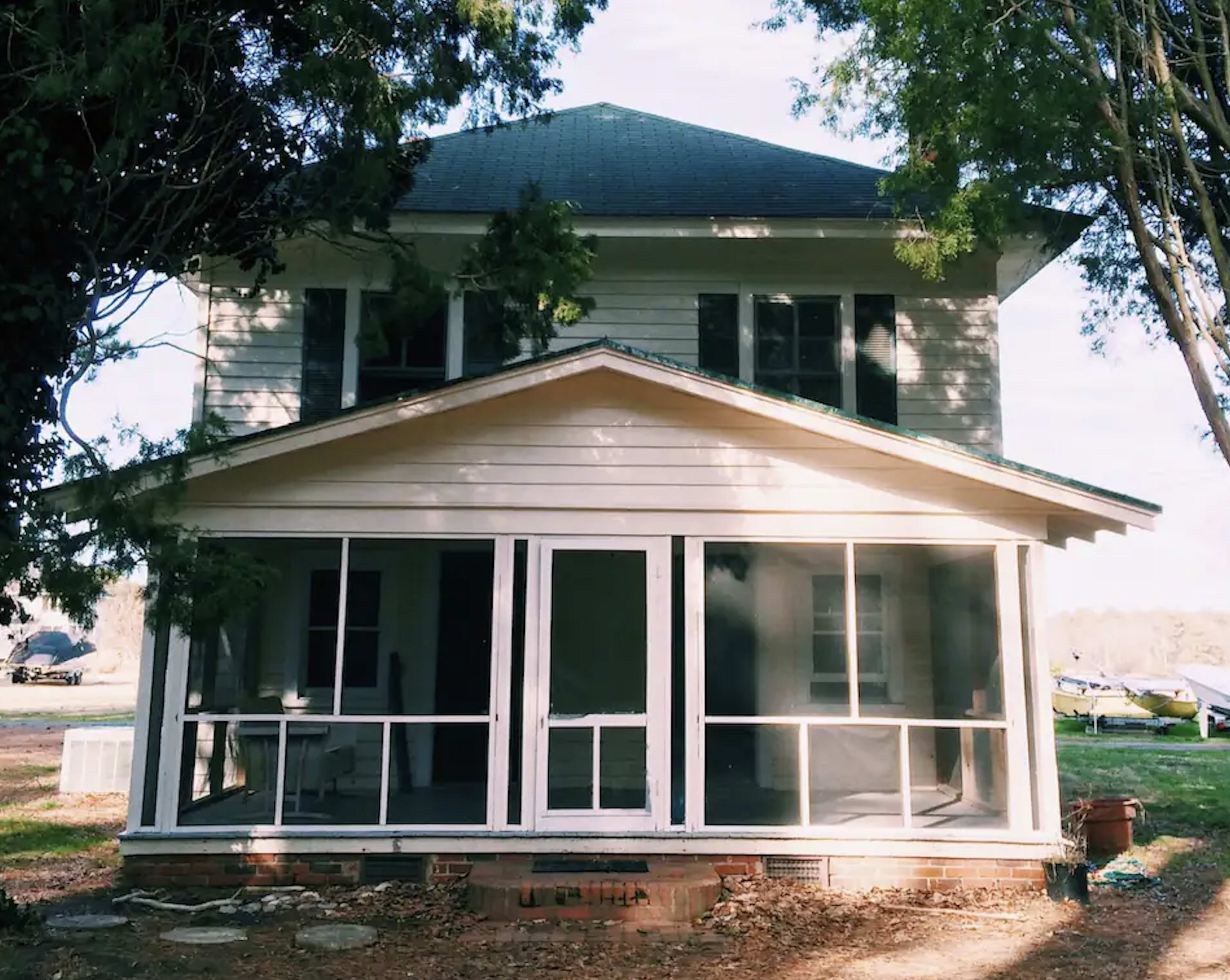 A two-story wooden house with a front porch featuring screens, surrounded by trees.