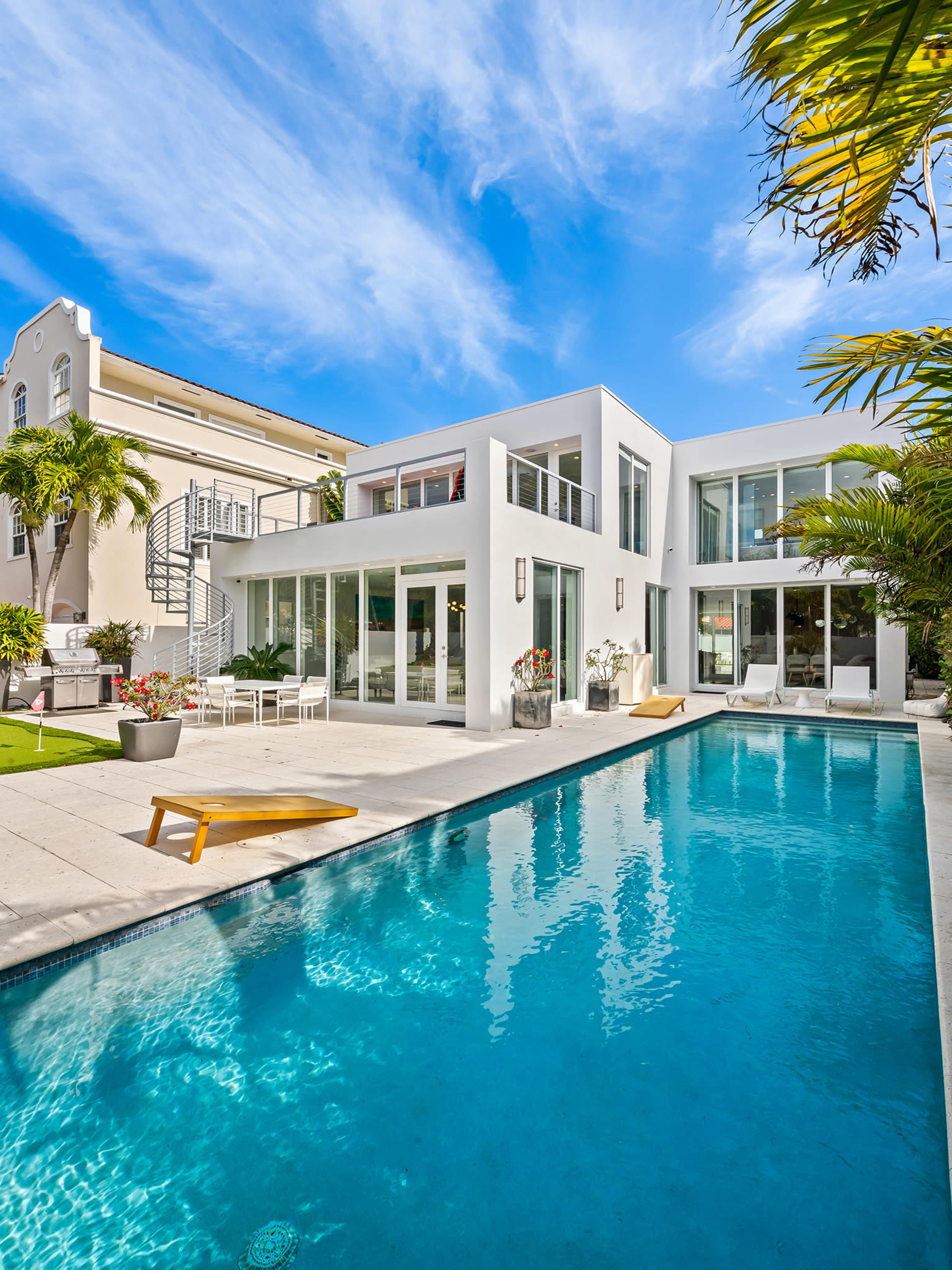 The image shows a modern, white house with large windows surrounding a swimming pool, complemented by tropical landscaping and clear blue skies.