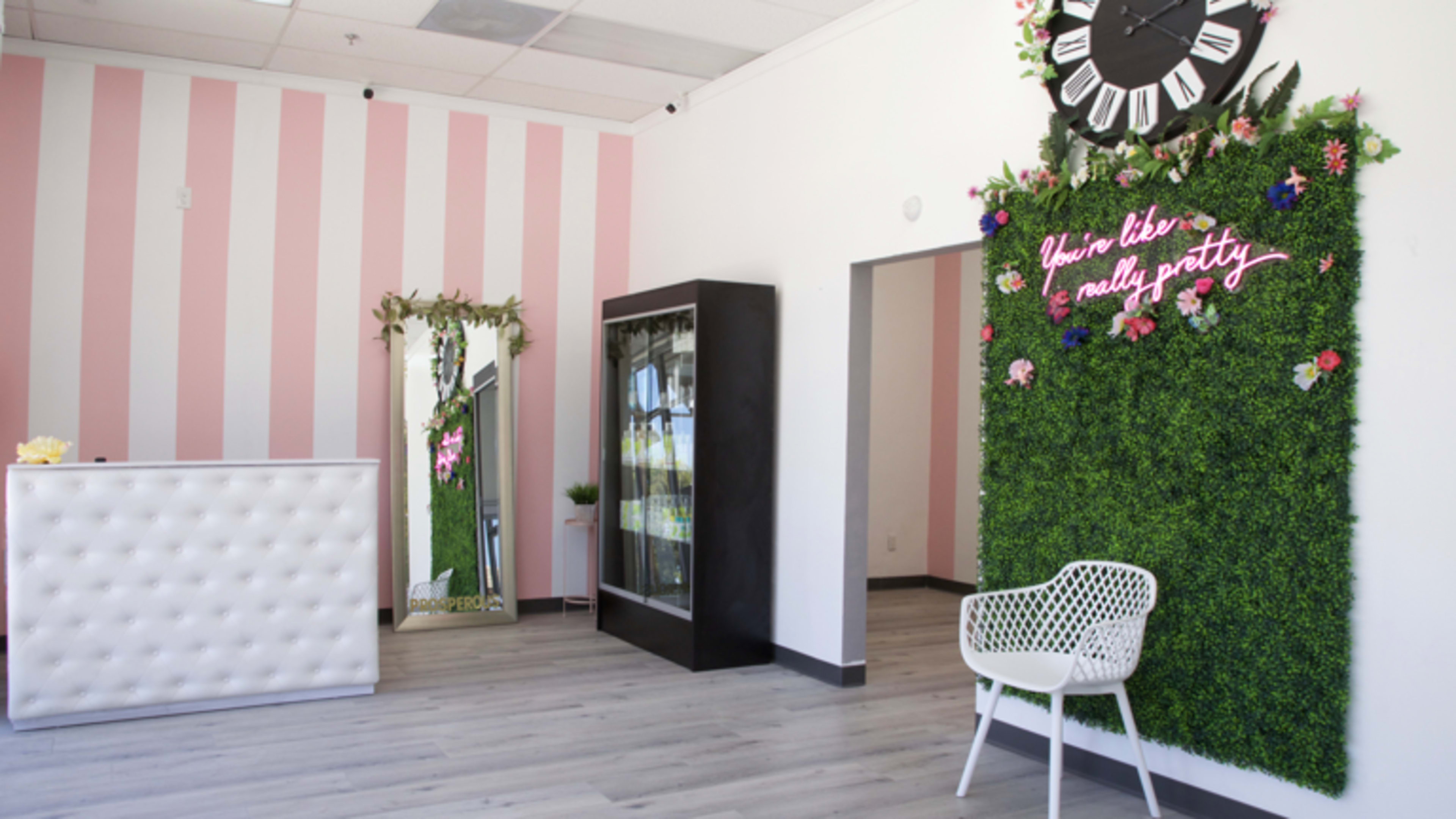The image shows a bright, modern reception area with pink striped walls, a mirrored wall, a black display refrigerator, and a decorative green wall with flowers and a clock.