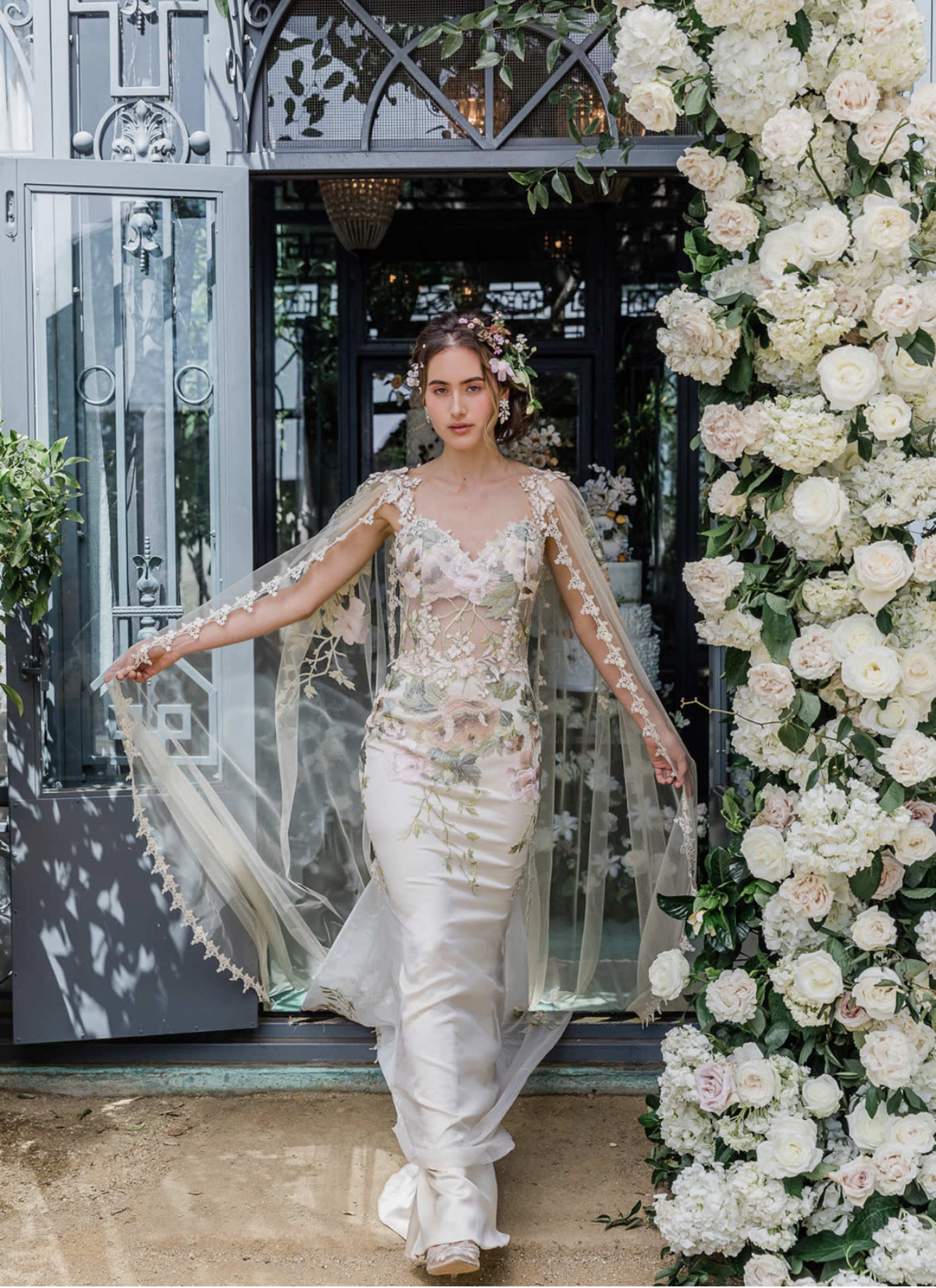 A bride in an intricate wedding gown with floral embellishments walks through a doorway adorned with greenery and white flowers.