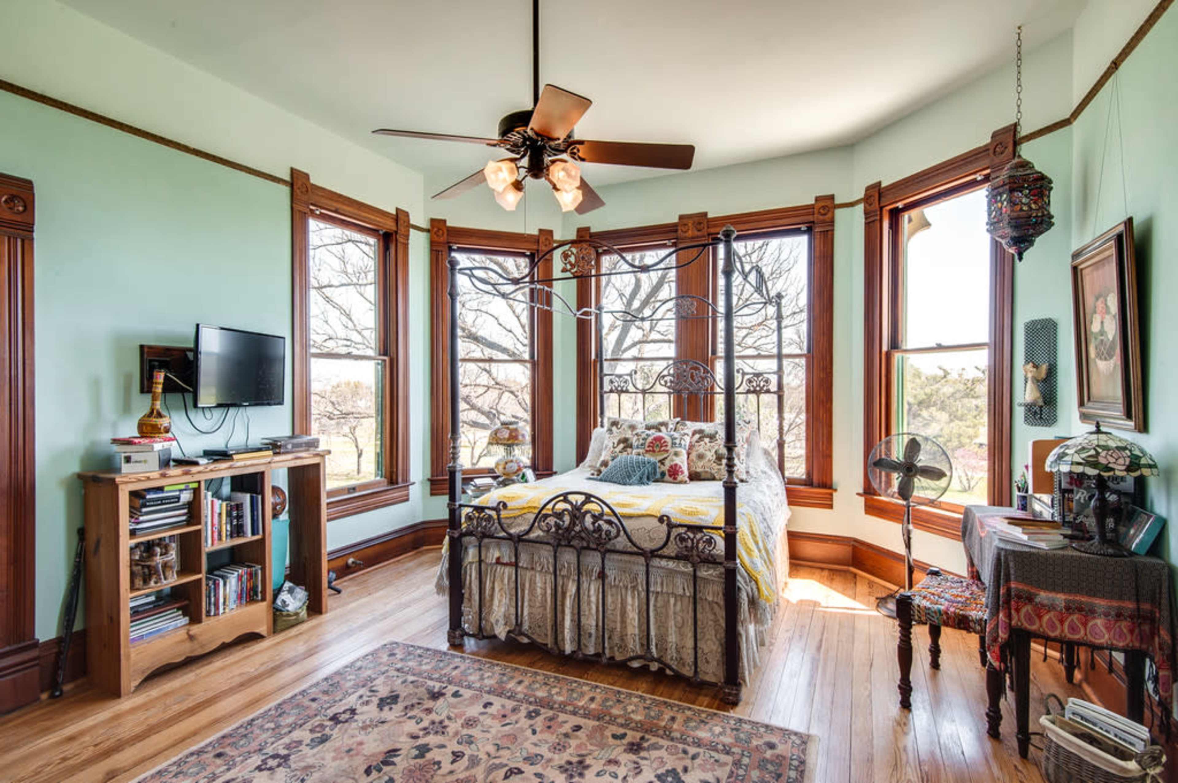 The image shows a bedroom with a large bed, a wooden bookshelf, a television mounted on the wall, and multiple windows providing natural light.