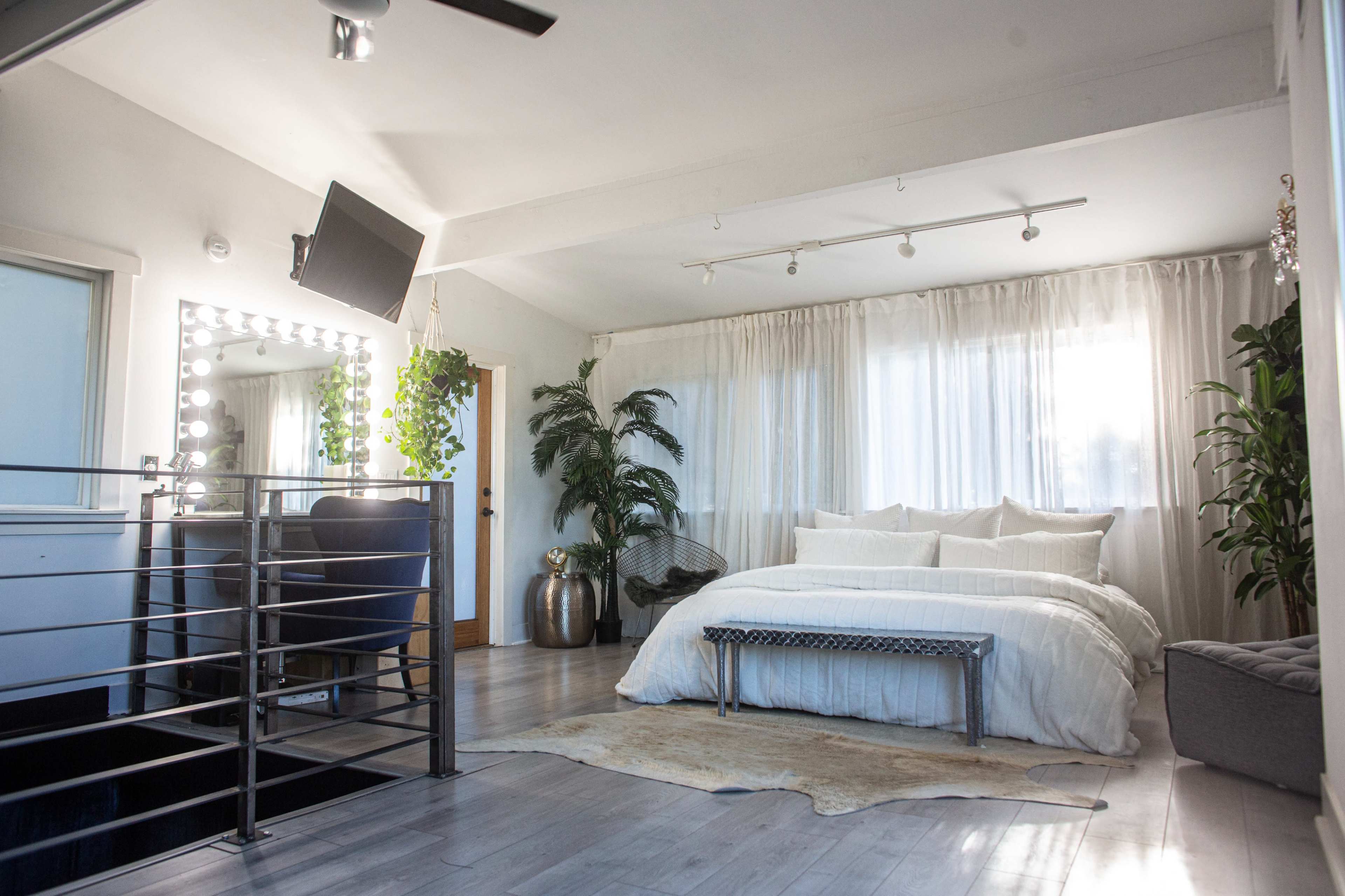 A spacious bedroom featuring a large bed with white linens, a mirror-lined vanity area, and plants placed throughout the room.