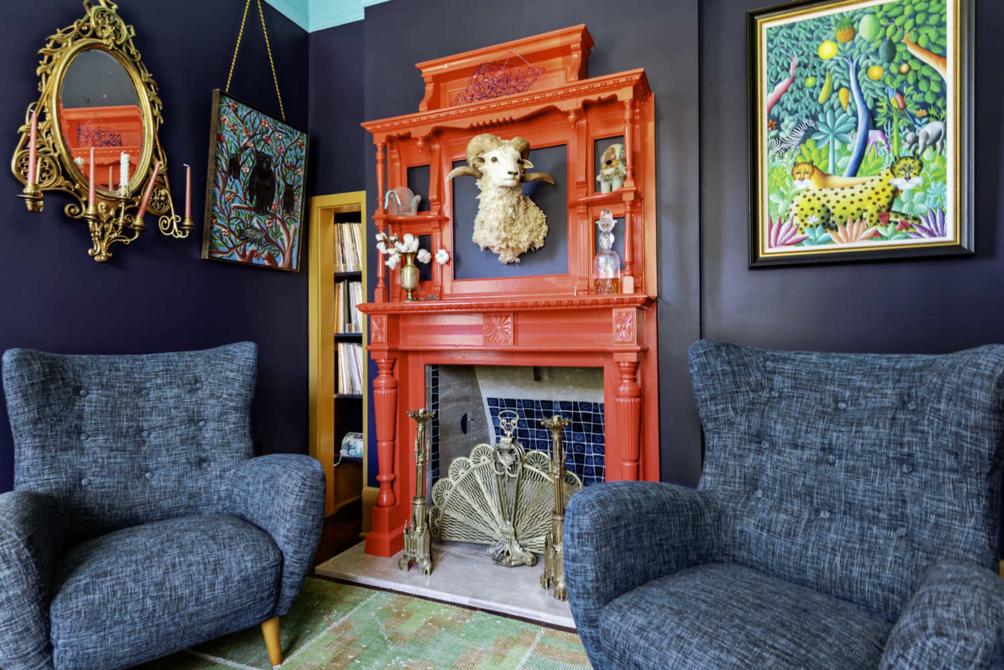 The image shows a colorful living room featuring an ornate orange fireplace adorned with decorative items, flanked by two textured gray armchairs and framed artwork on the walls.