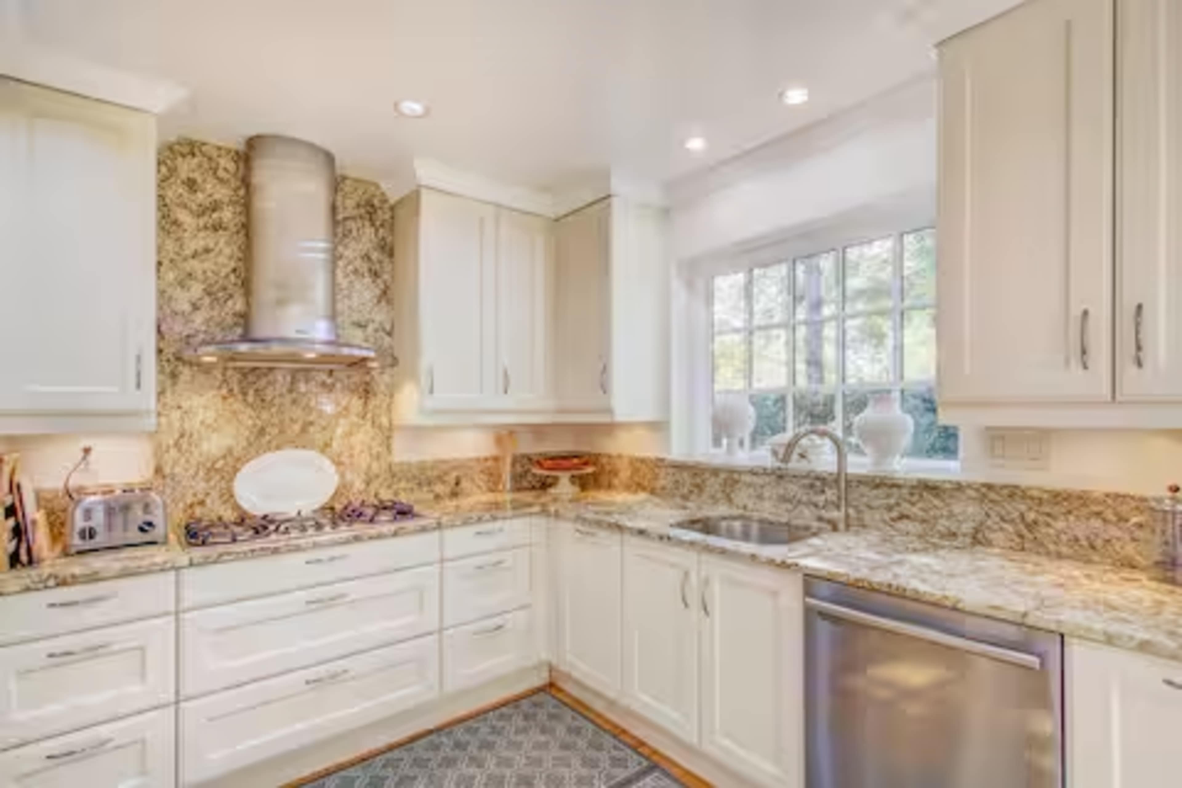 The image shows a modern kitchen featuring white cabinetry, a granite countertop, and a large window over the sink.