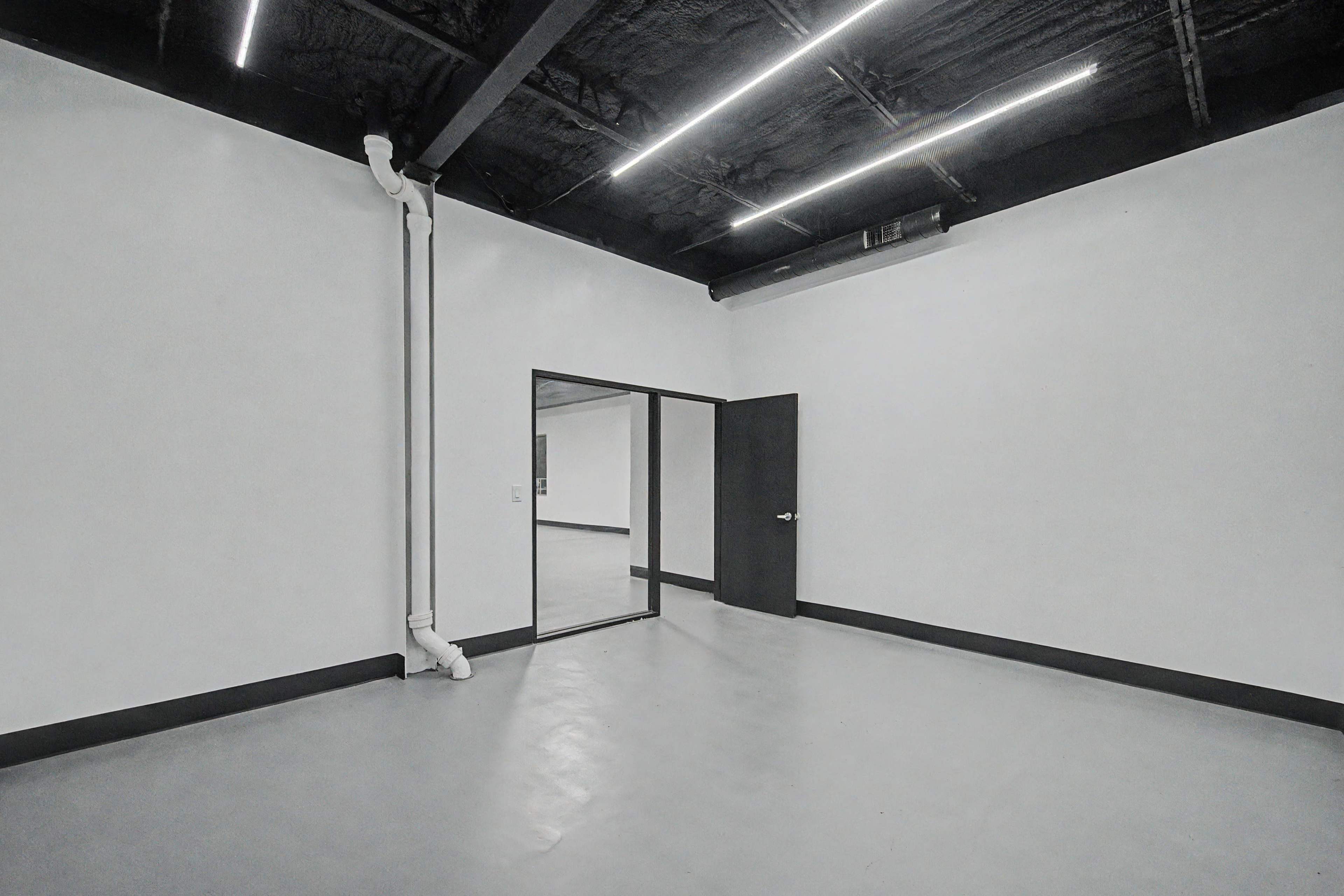 The image shows an empty room with plain white walls, gray flooring, and a doorway leading to another space.