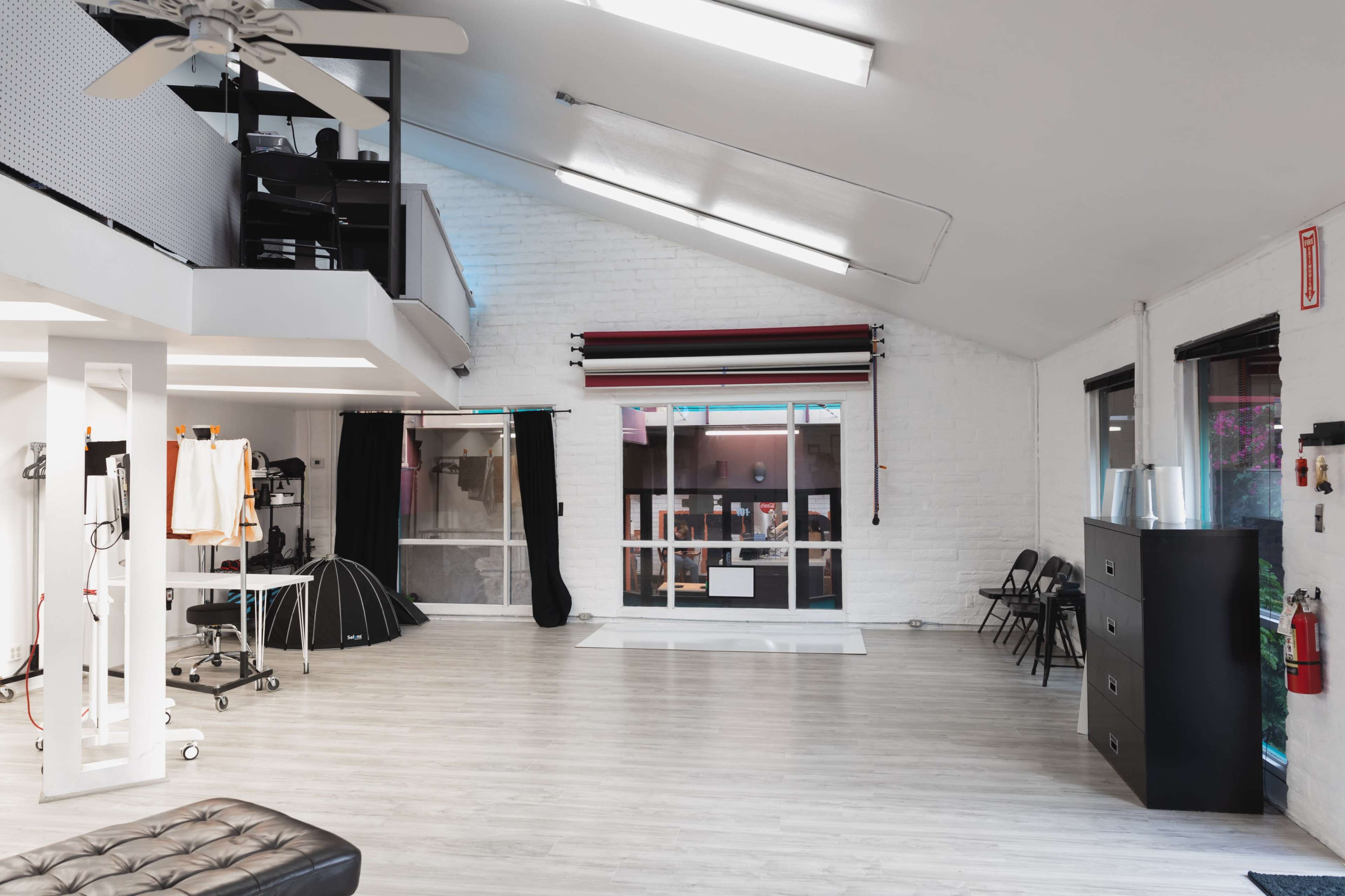 A spacious, well-lit studio with white walls, wooden flooring, and large windows, featuring various furniture and equipment along the sides.