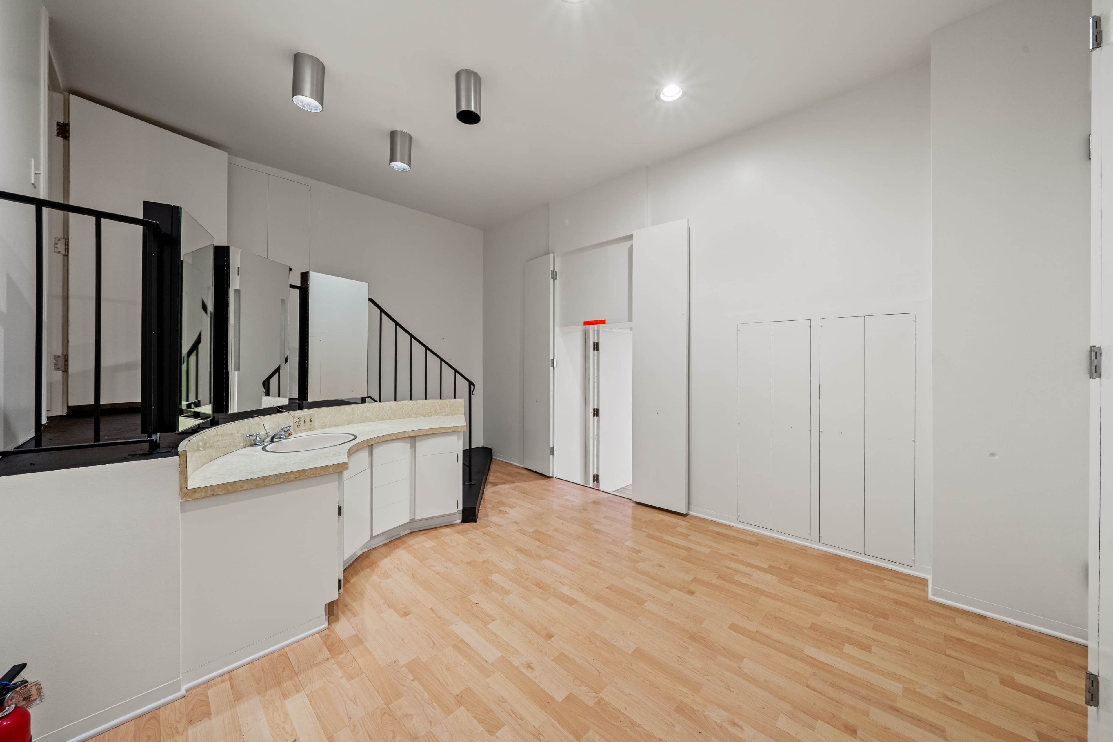 The image depicts a spacious interior room with light wood flooring, a curved countertop, and a staircase leading to an upper level.