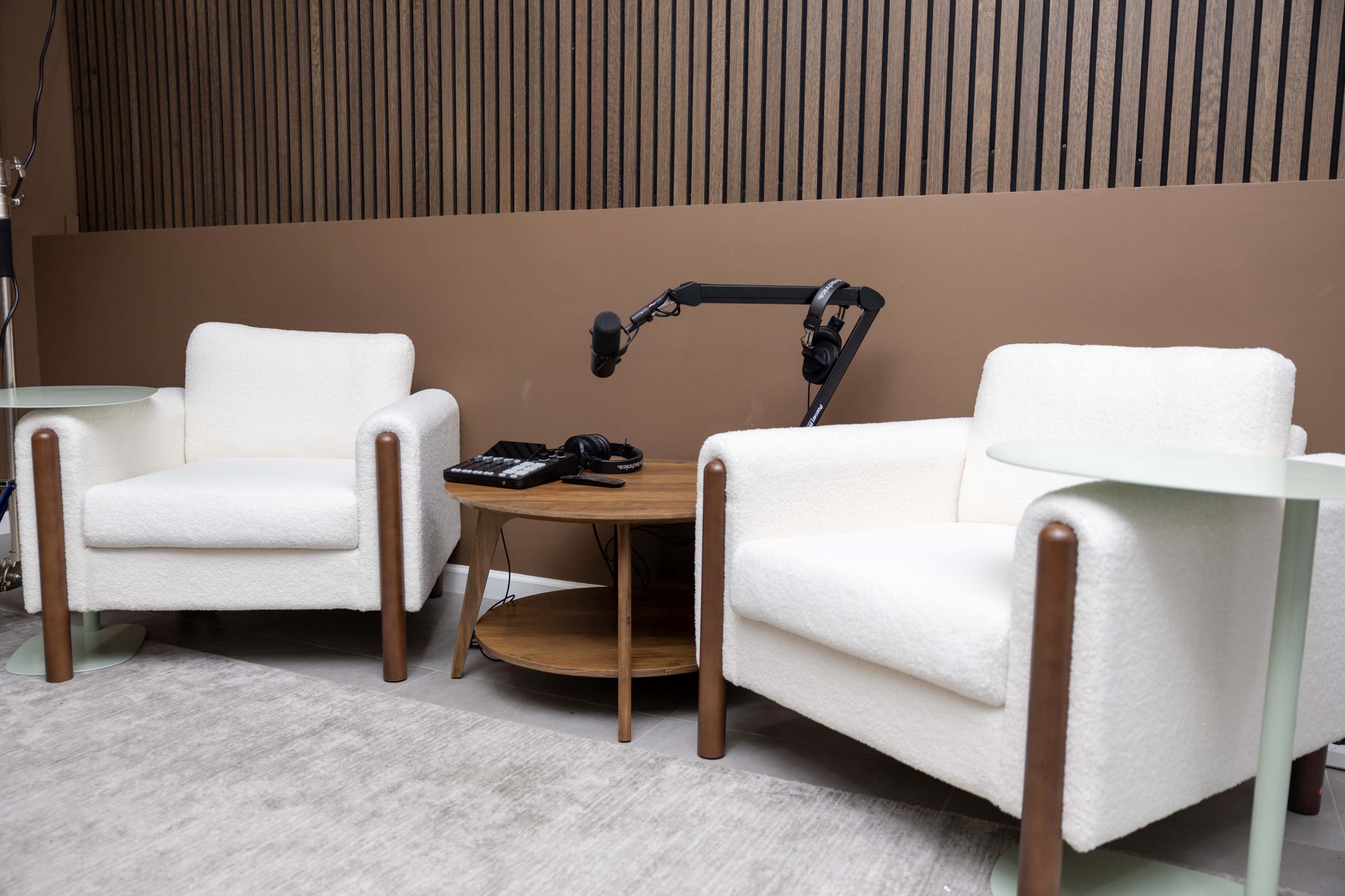 The image shows two white armchairs beside a wooden side table with a microphone mounted on an arm and a pair of headphones on top.