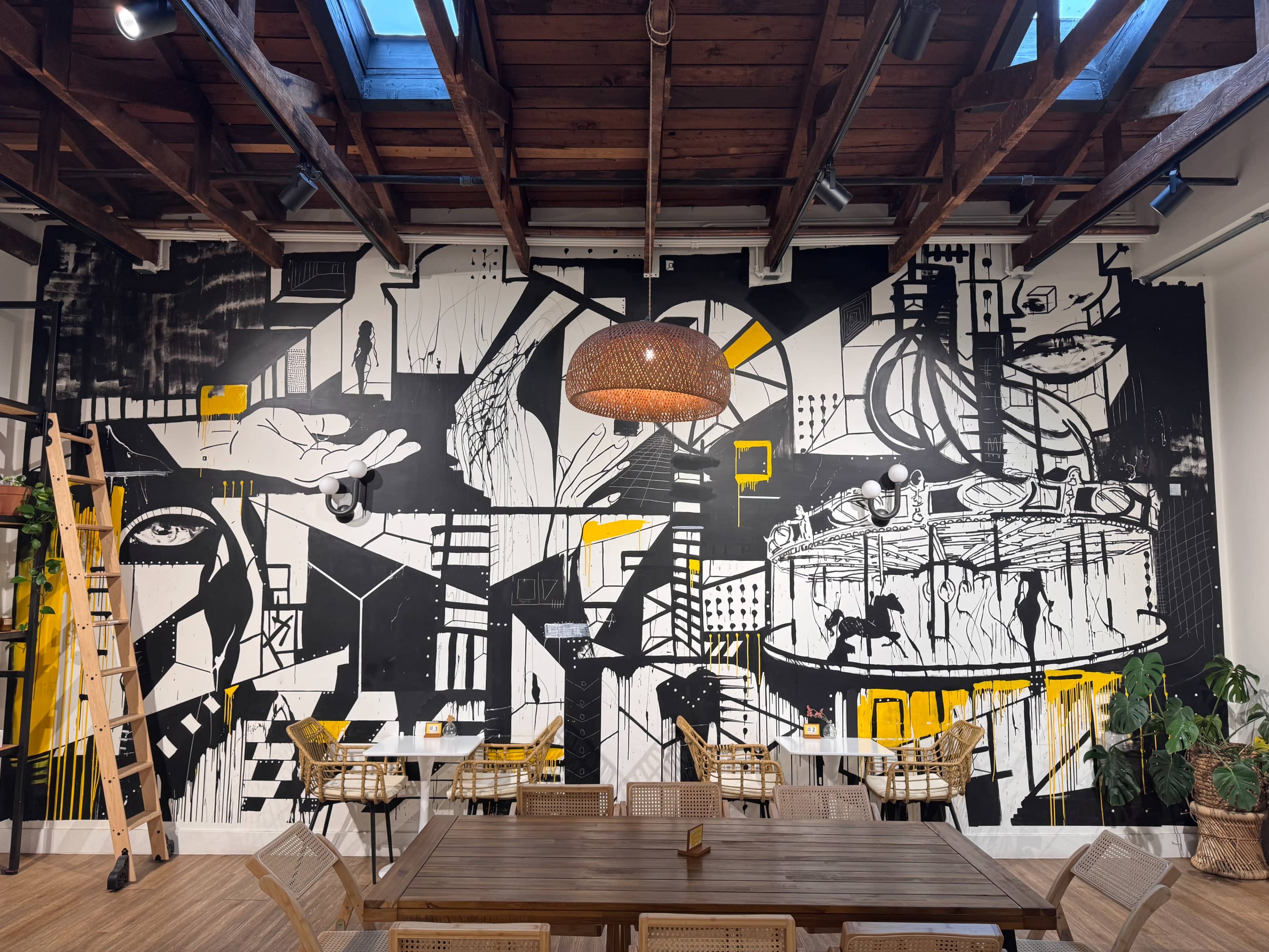 The image shows a large black and white mural with abstract shapes and figures, featuring yellow accents, on a wall of a cafe or common area with wooden beams and a central light fixture.