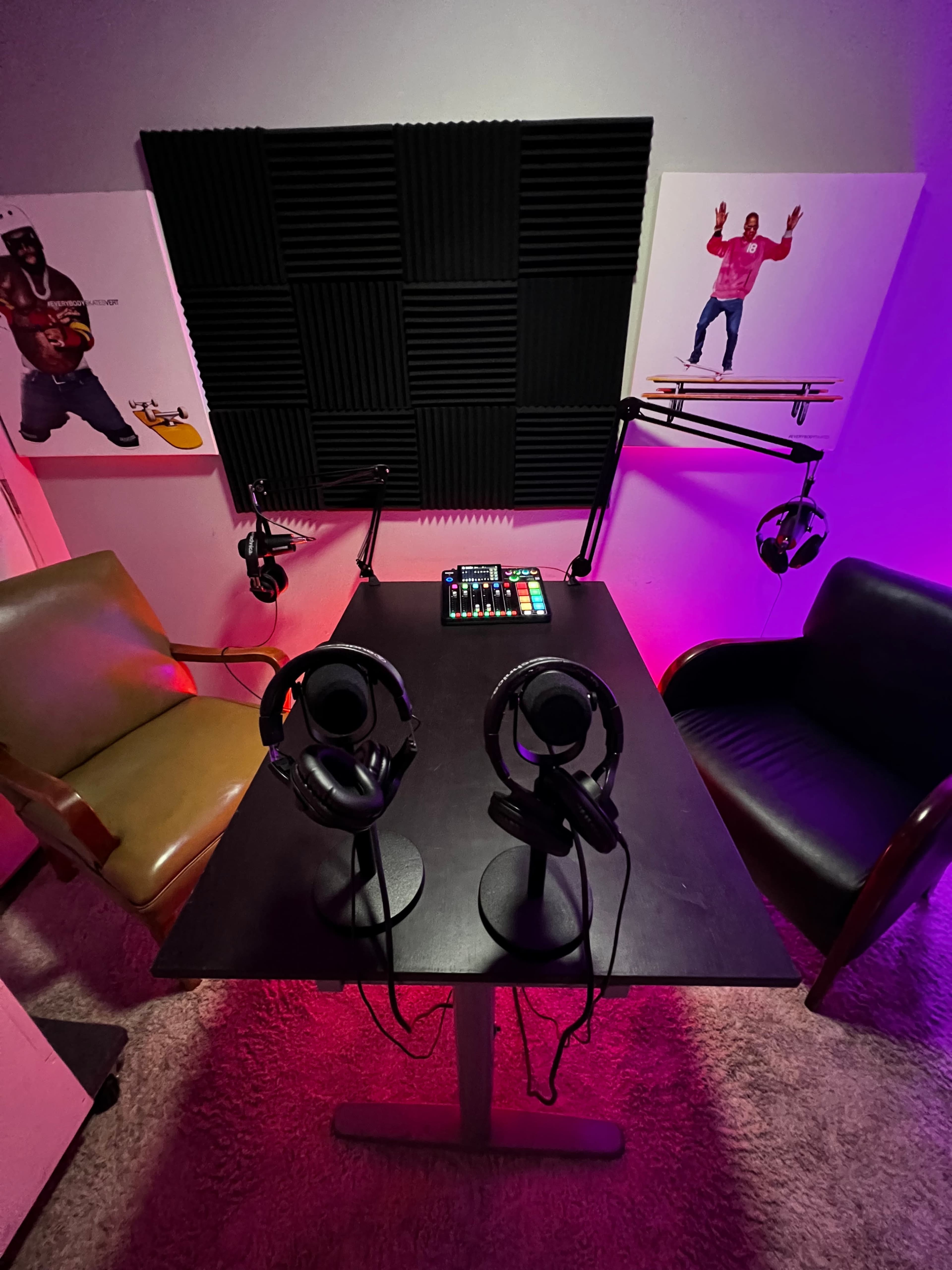 A podcast recording setup with two chairs, a table, headphones on stands, and a control panel, surrounded by acoustic panels on the walls and ambient lighting.