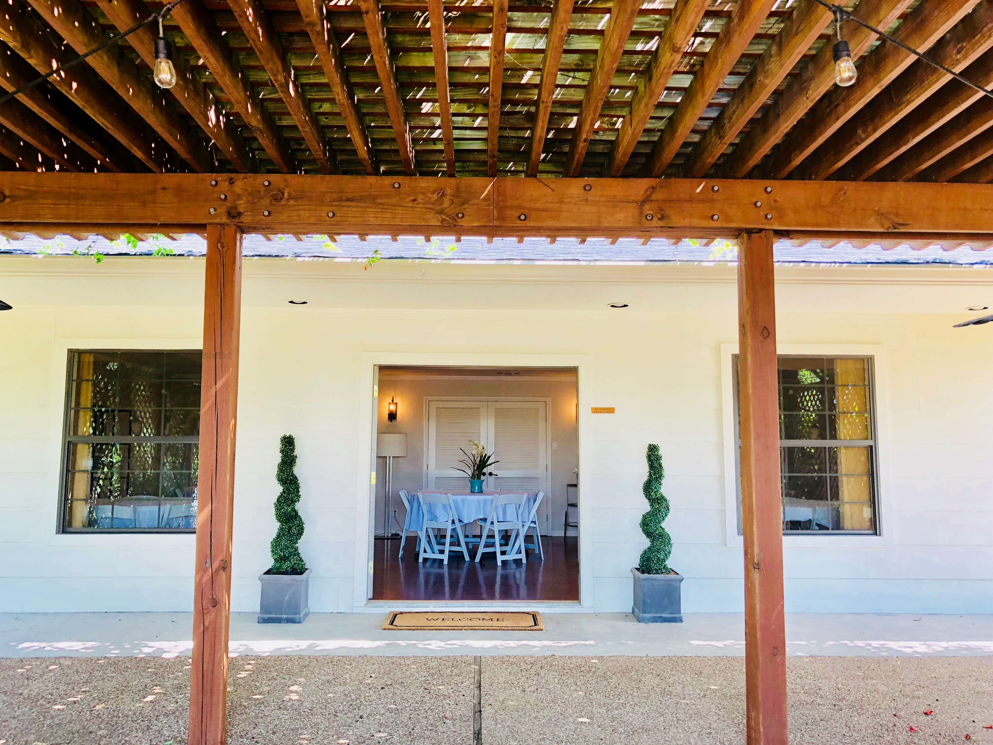 A covered porch with wooden beams leads to a white entrance featuring two potted topiary plants on either side.