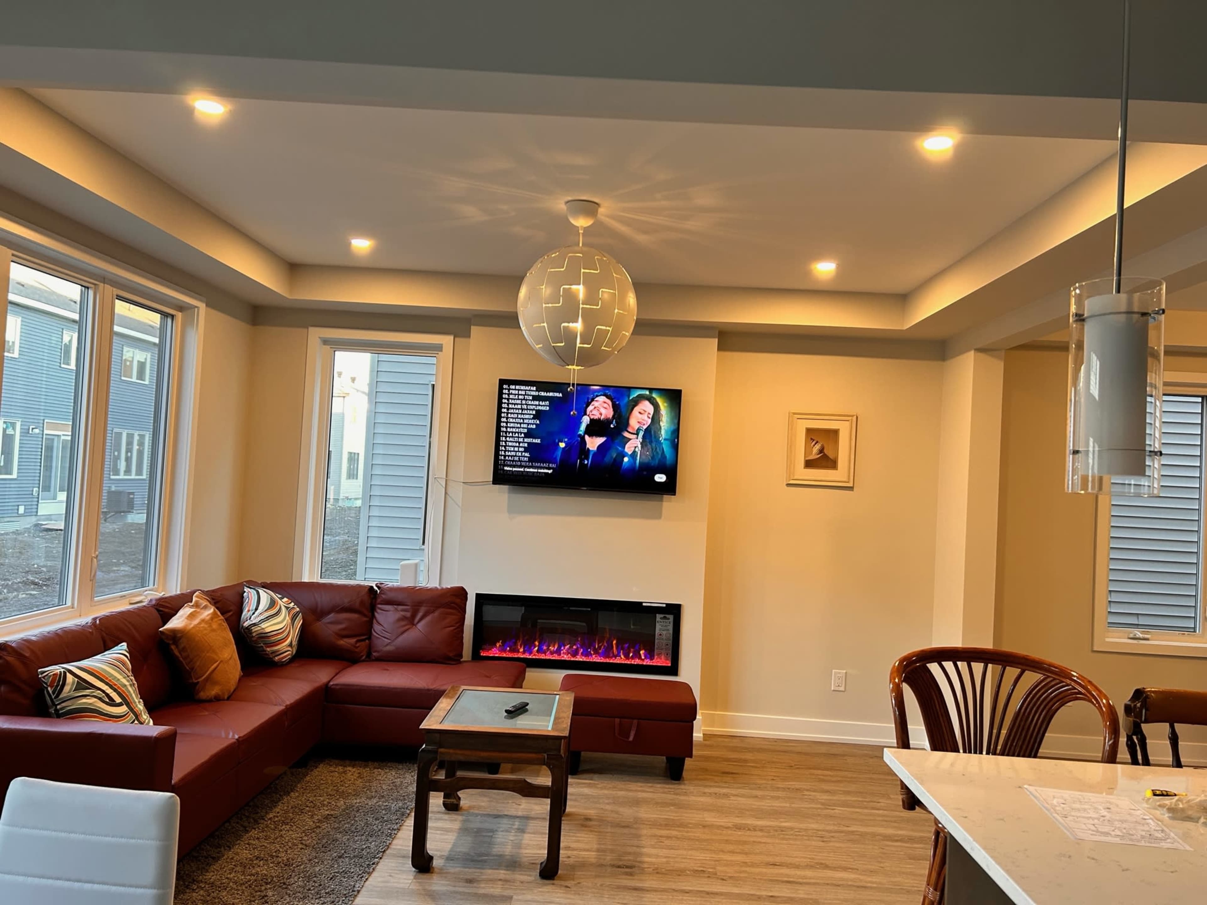 A modern living room features a leather sofa, a wall-mounted television displaying a performance, and an electric fireplace underneath.