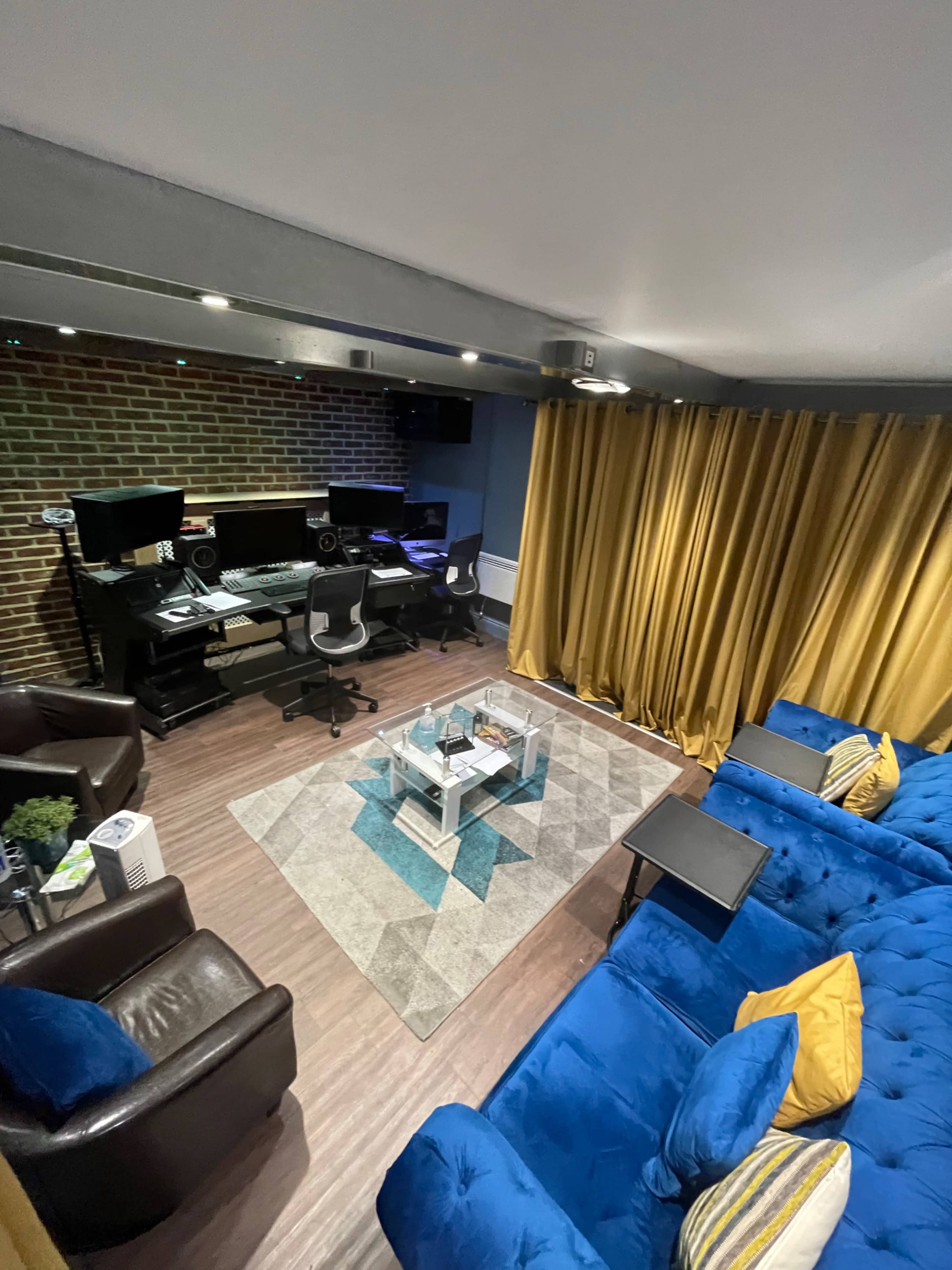A music studio featuring two computer workstations, a blue sofa with yellow cushions, brown chairs, and a patterned rug, all set against a brick wall with gold curtains.