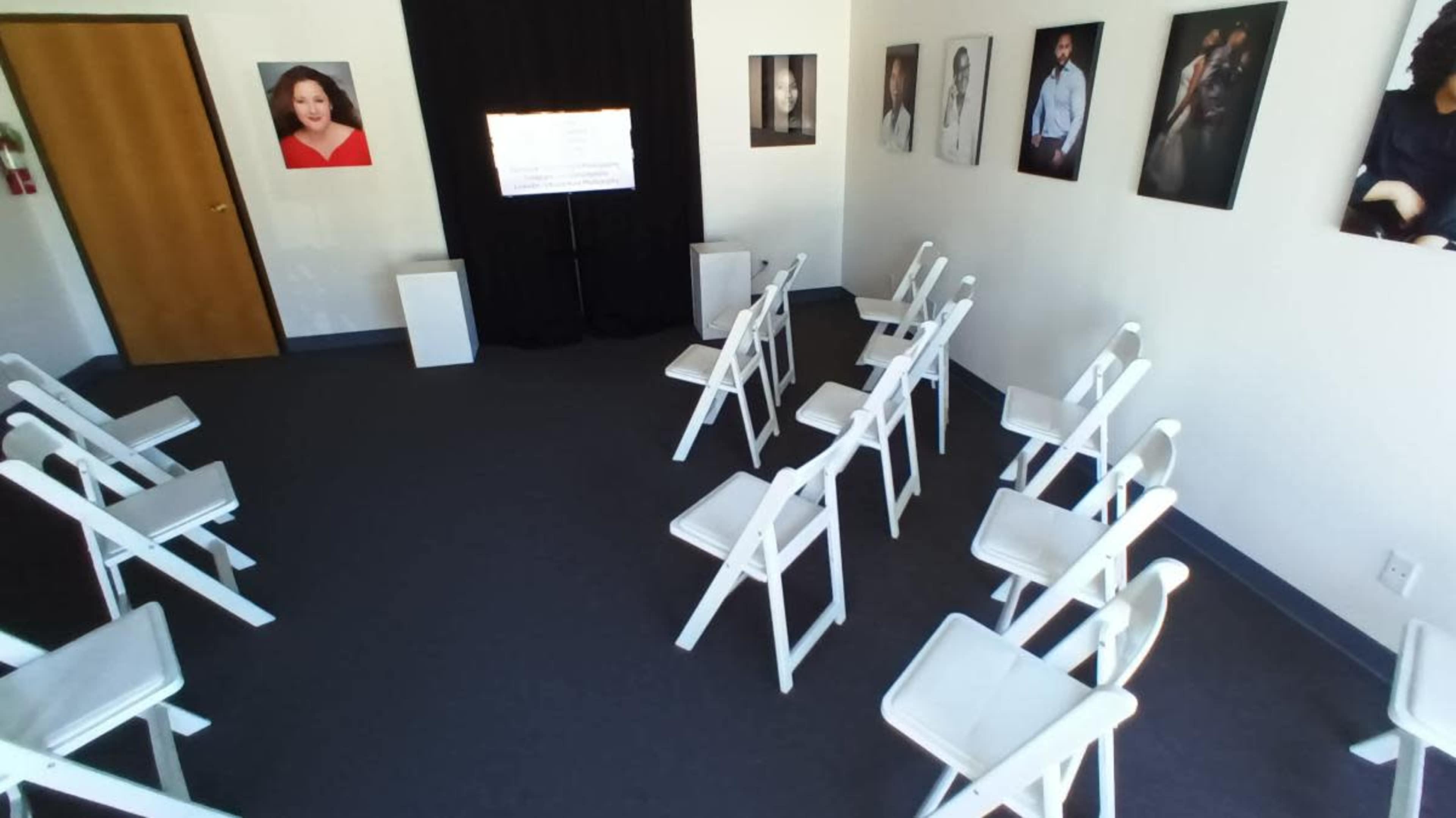 The image shows a small, empty room arranged with white folding chairs facing a screen, surrounded by framed photographs on the walls.