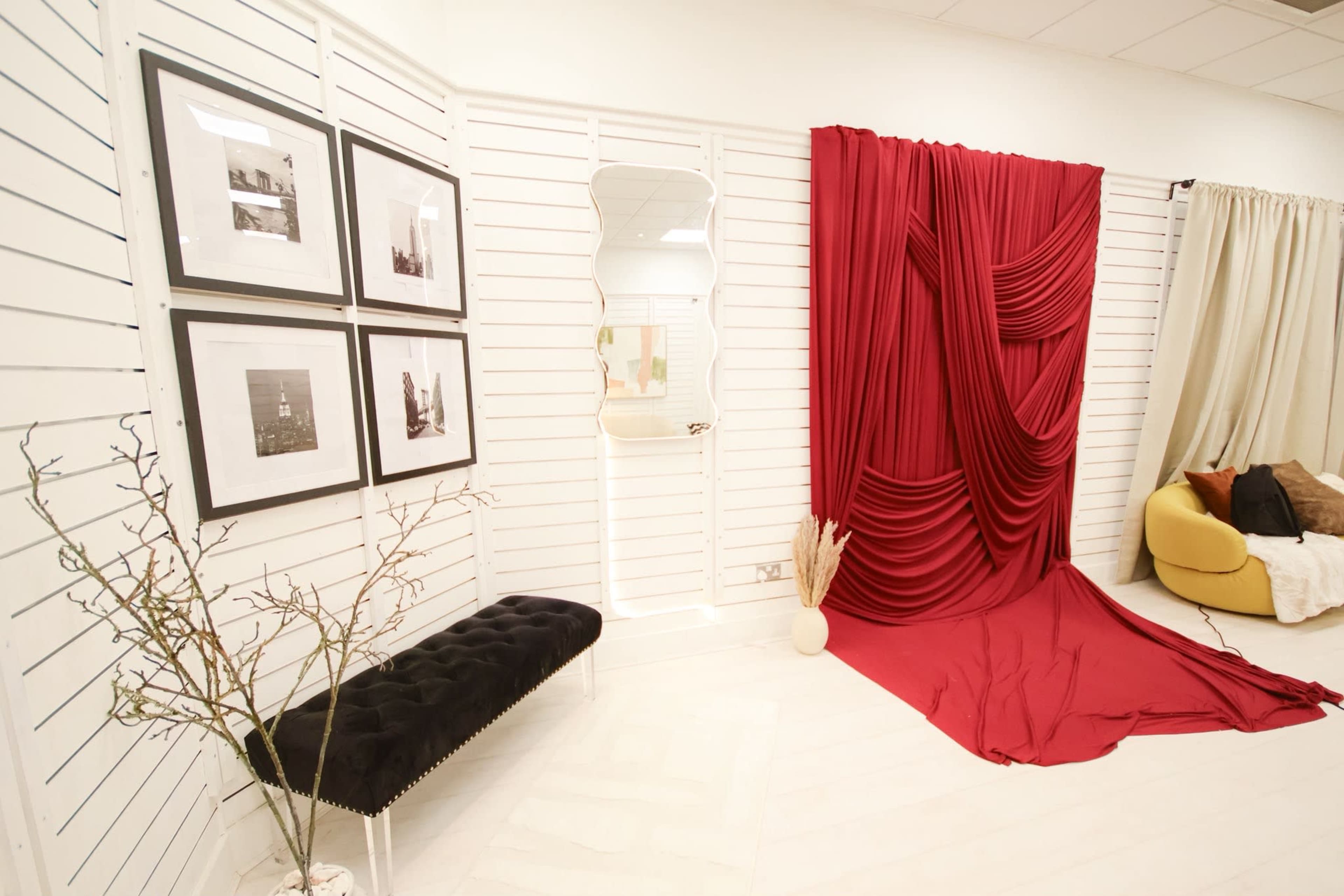 The image shows a decorated interior space featuring a red draped curtain, framed photographs on the wall, a mirror, a black bench, and a yellow sofa.