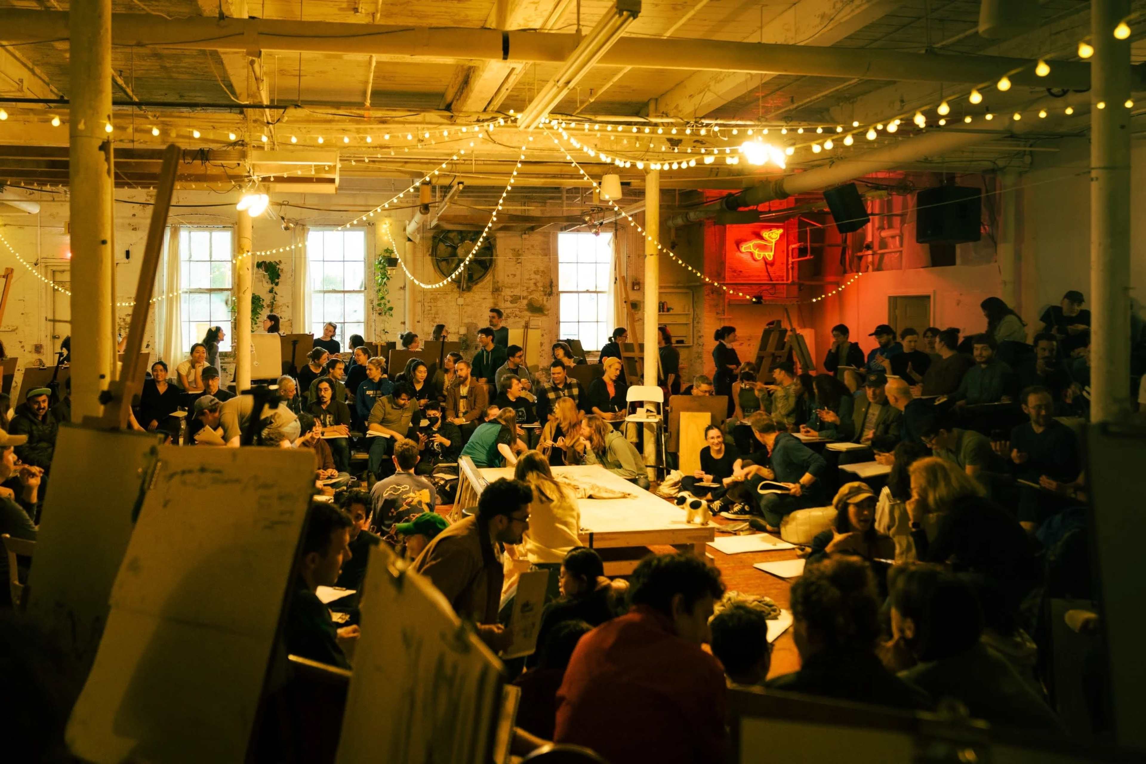 A crowded art studio is filled with people engaged in drawing and sketching activities, under warm lighting and string lights.