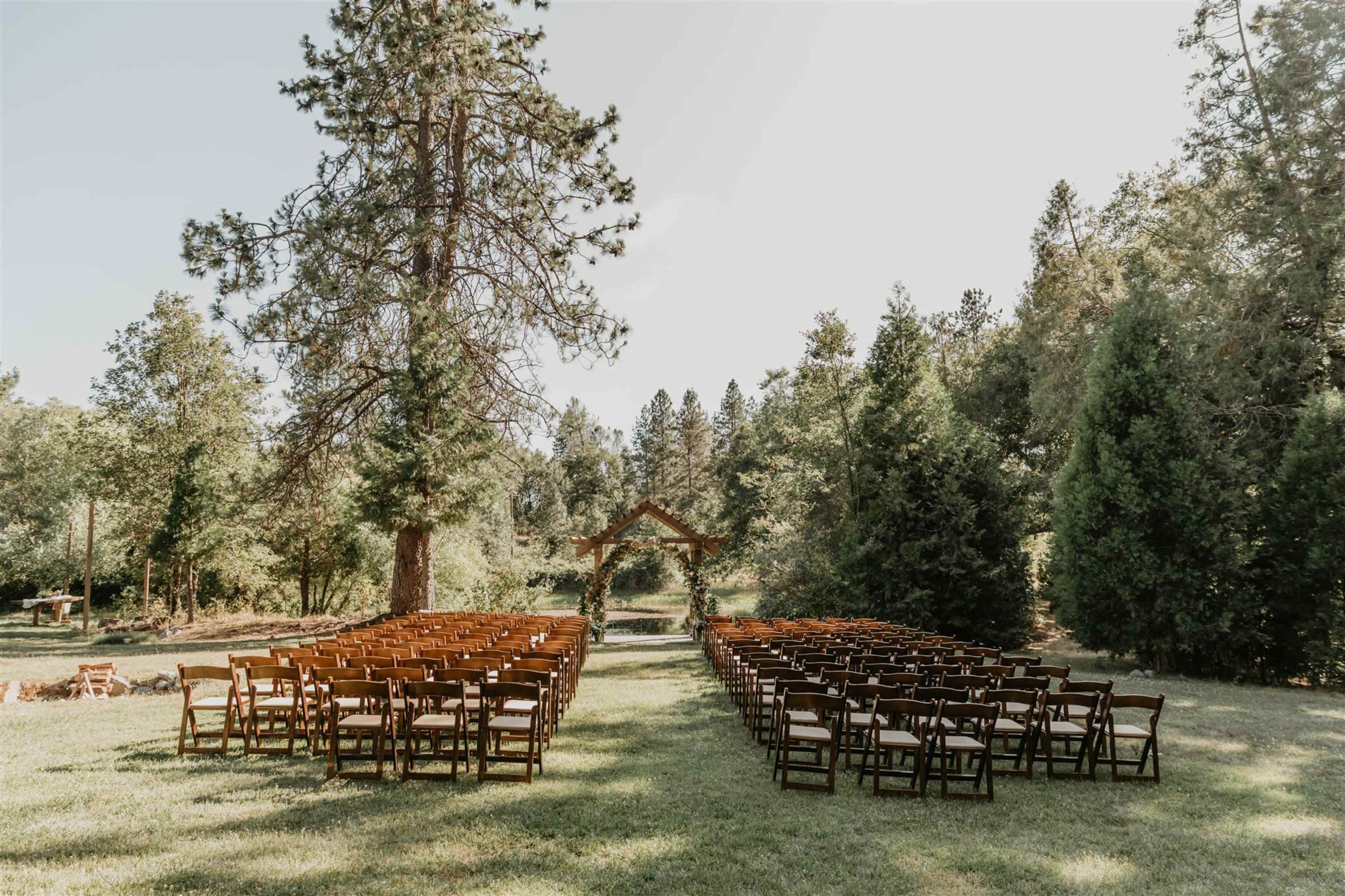The image shows a spacious outdoor venue set up for a wedding ceremony, with rows of wooden chairs facing a decorative arch under a large tree.