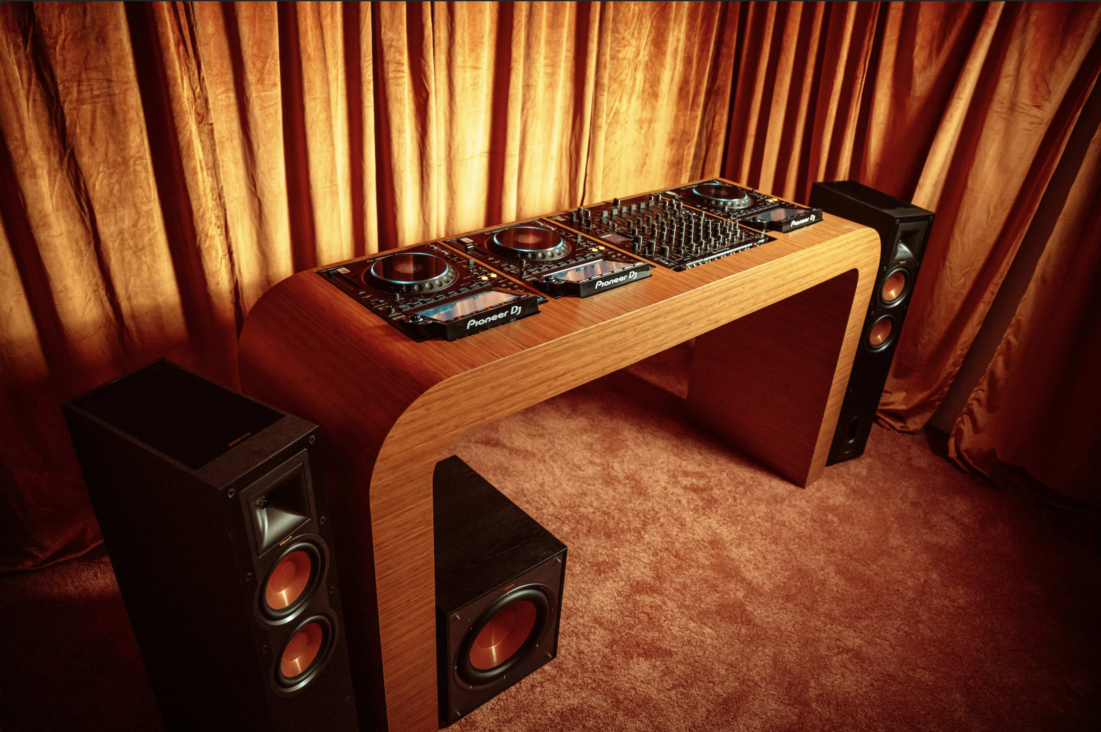 A wooden DJ table with Pioneer equipment is positioned between two large speakers in front of a curtain.