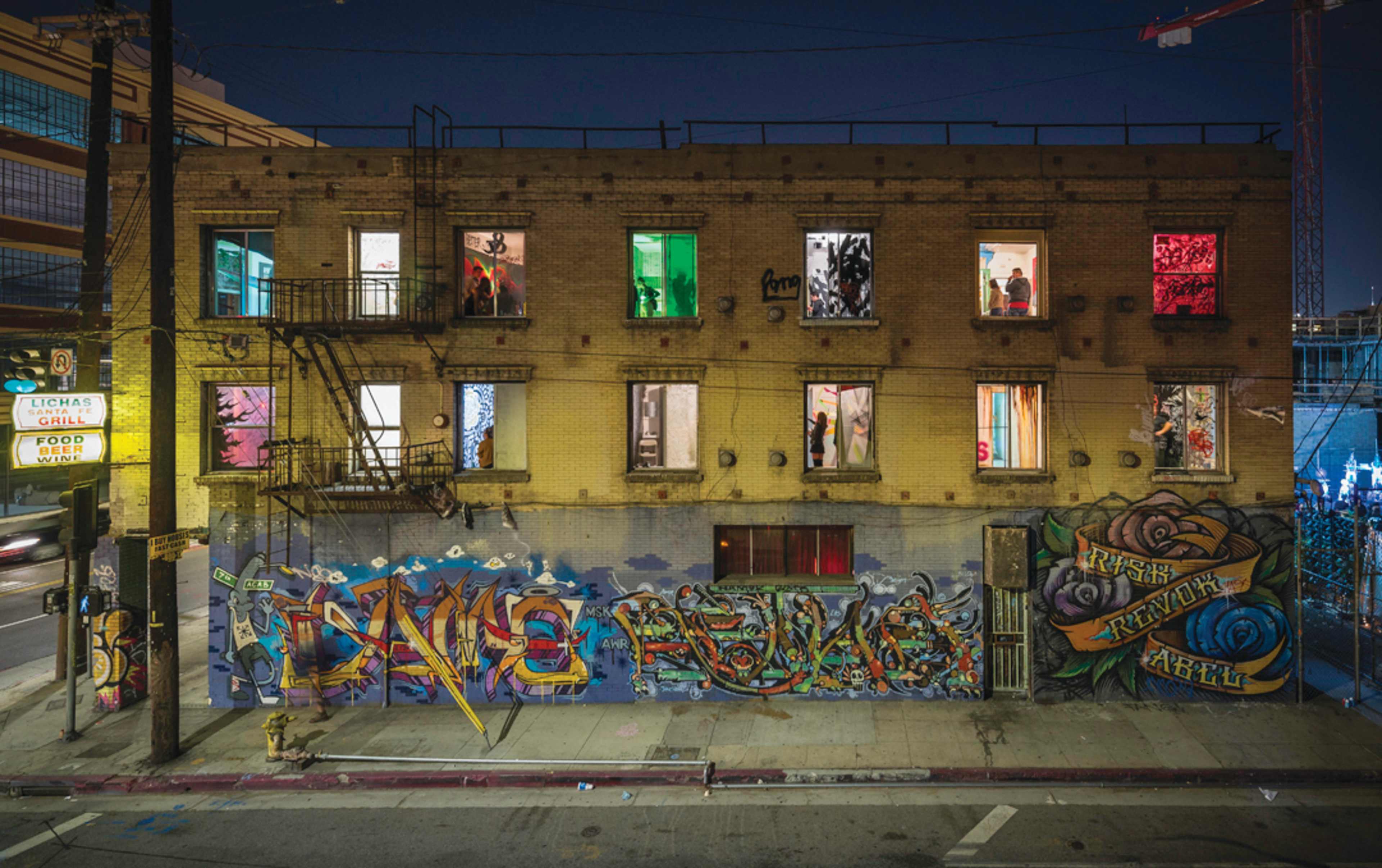 A brick building with several illuminated windows displays vibrant graffiti on its exterior and a mural covering the adjacent wall.