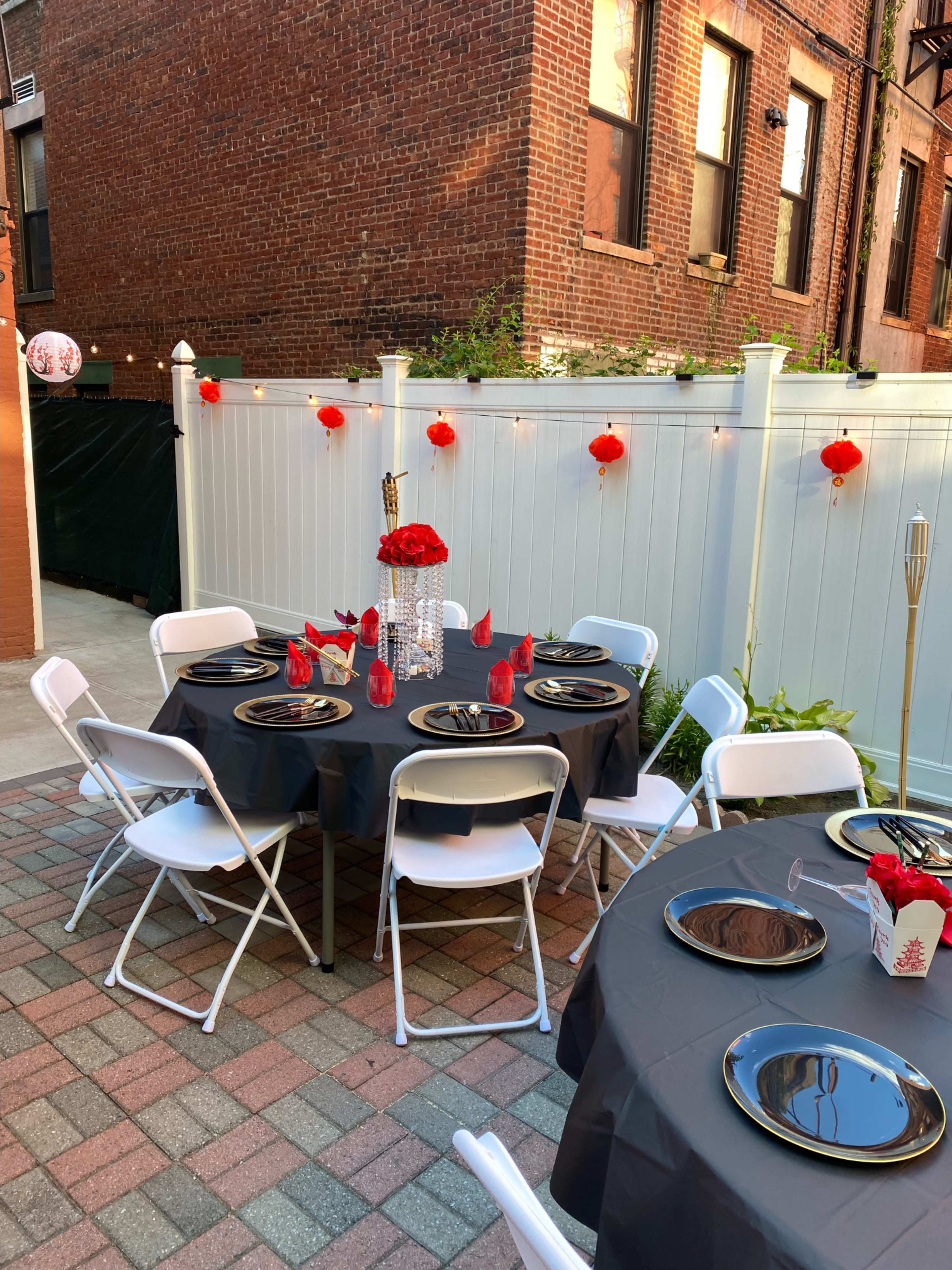 A set dining area features round tables with black tablecloths, adorned with red centerpieces and decorative lanterns, in a brick-walled courtyard.