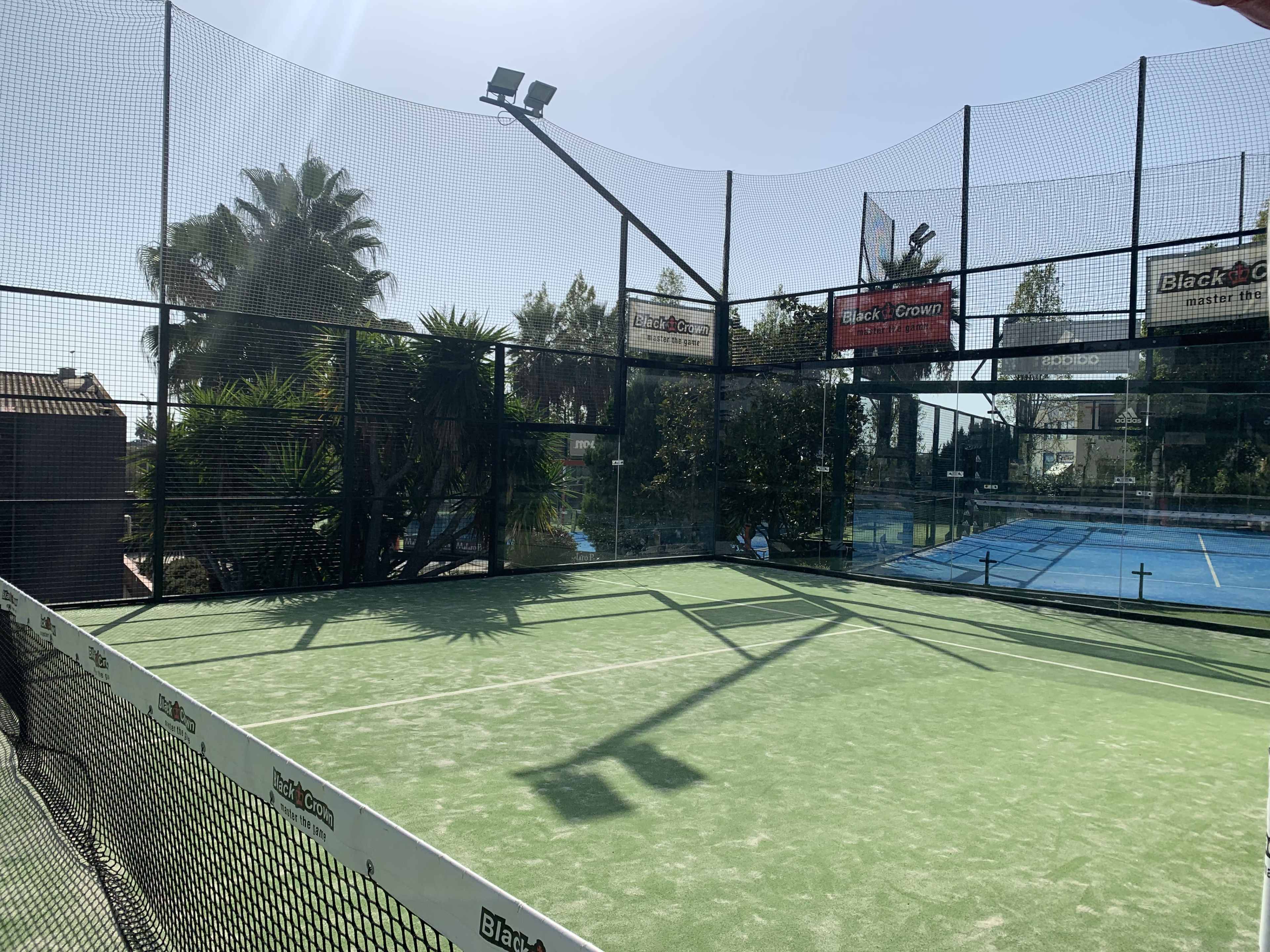 A well-maintained padel court surrounded by palm trees and a blue tennis court in the background.
