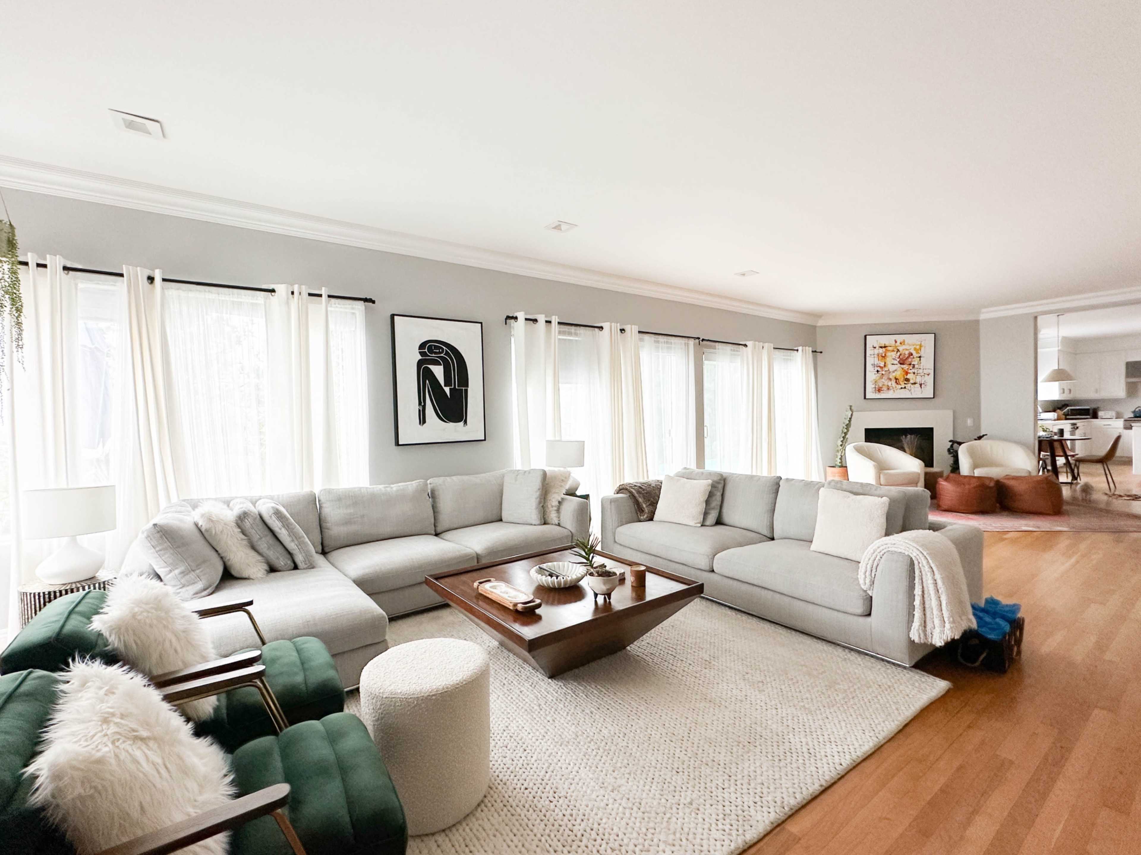 The image shows a spacious living room with a sectional sofa, large windows dressed in sheer curtains, and a wooden coffee table at the center.