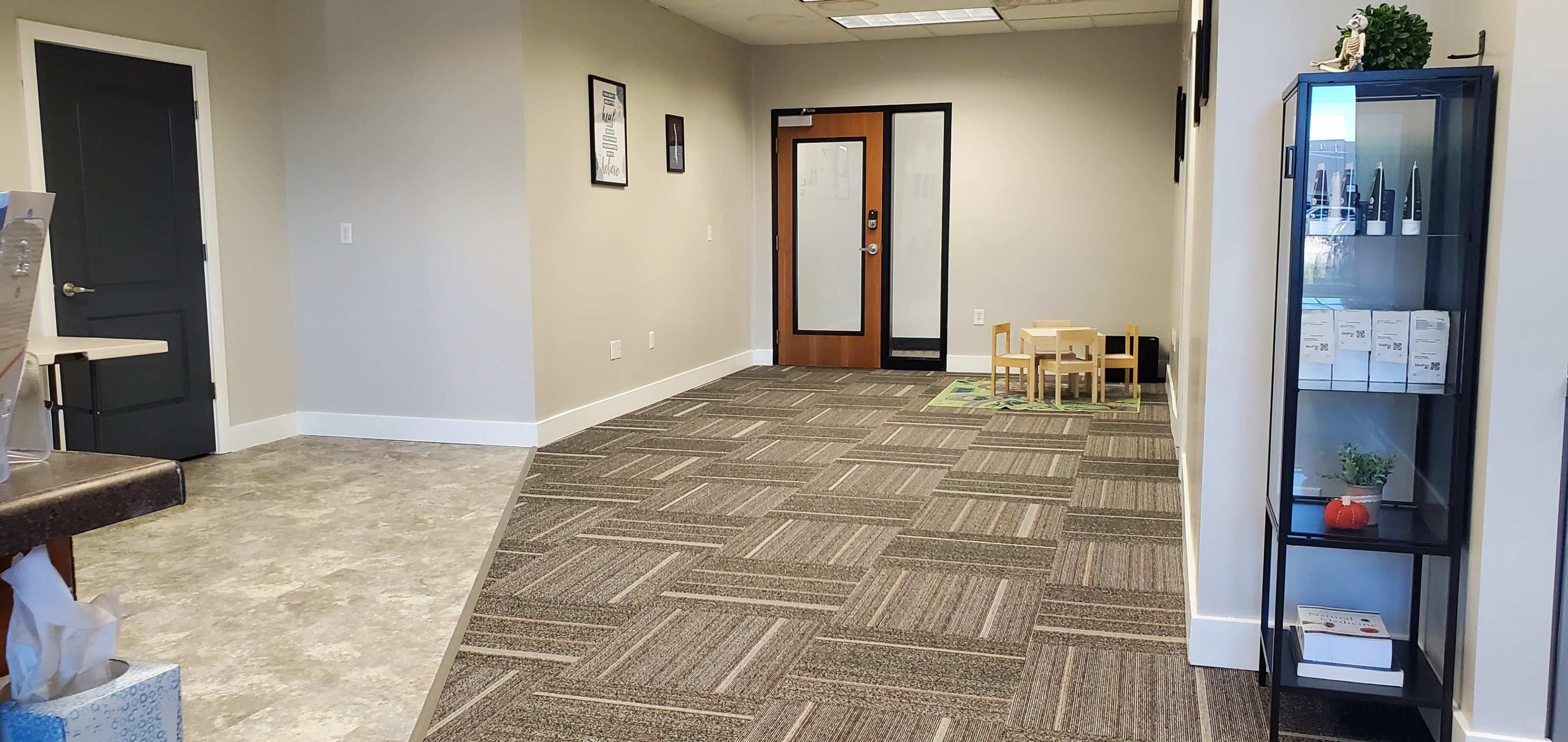 A clean, modern entryway with gray carpet, a wooden door, and a small play area with child-sized furniture in a well-lit space.