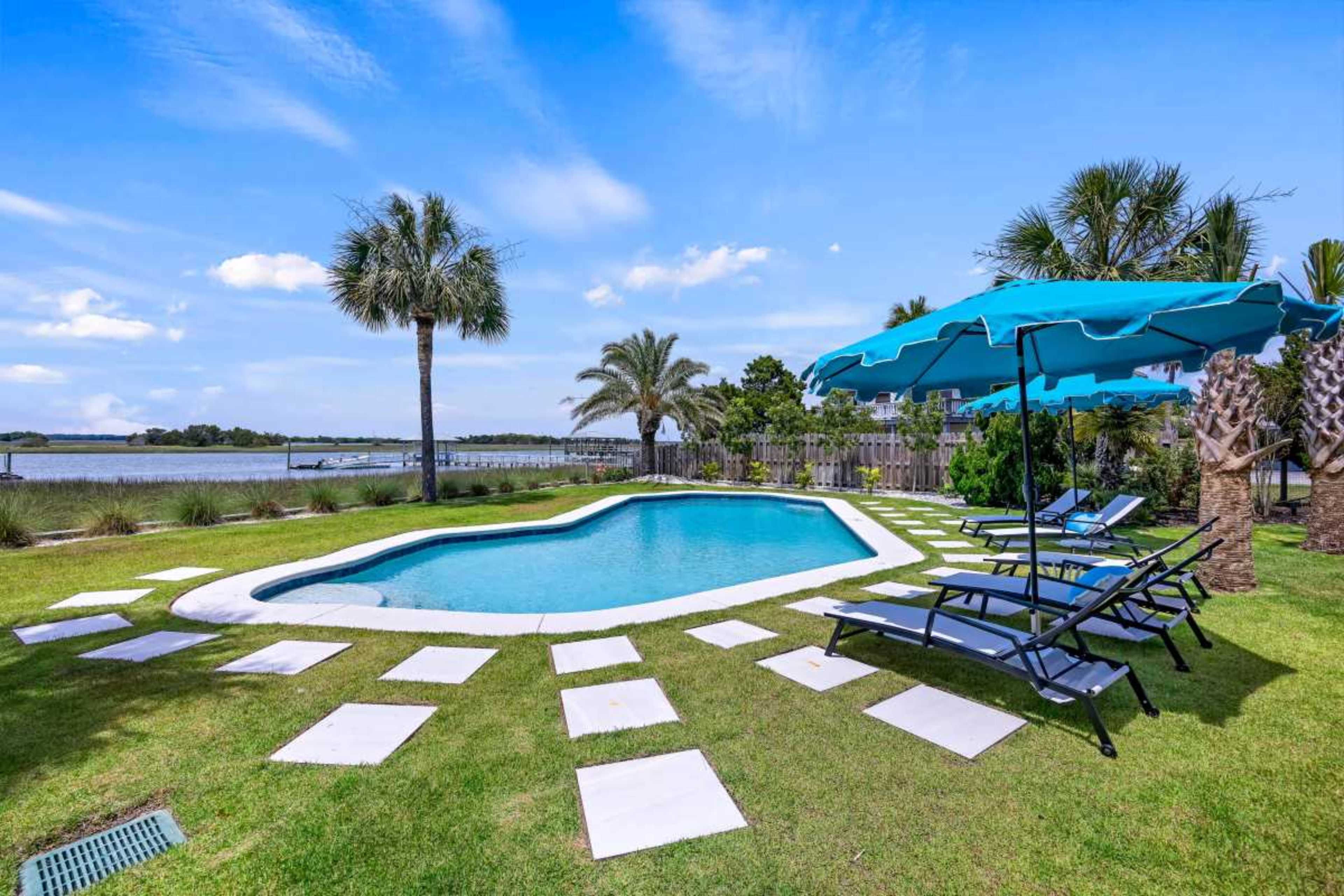 The image shows a swimming pool surrounded by lounge chairs and palm trees, with a view of water in the background.