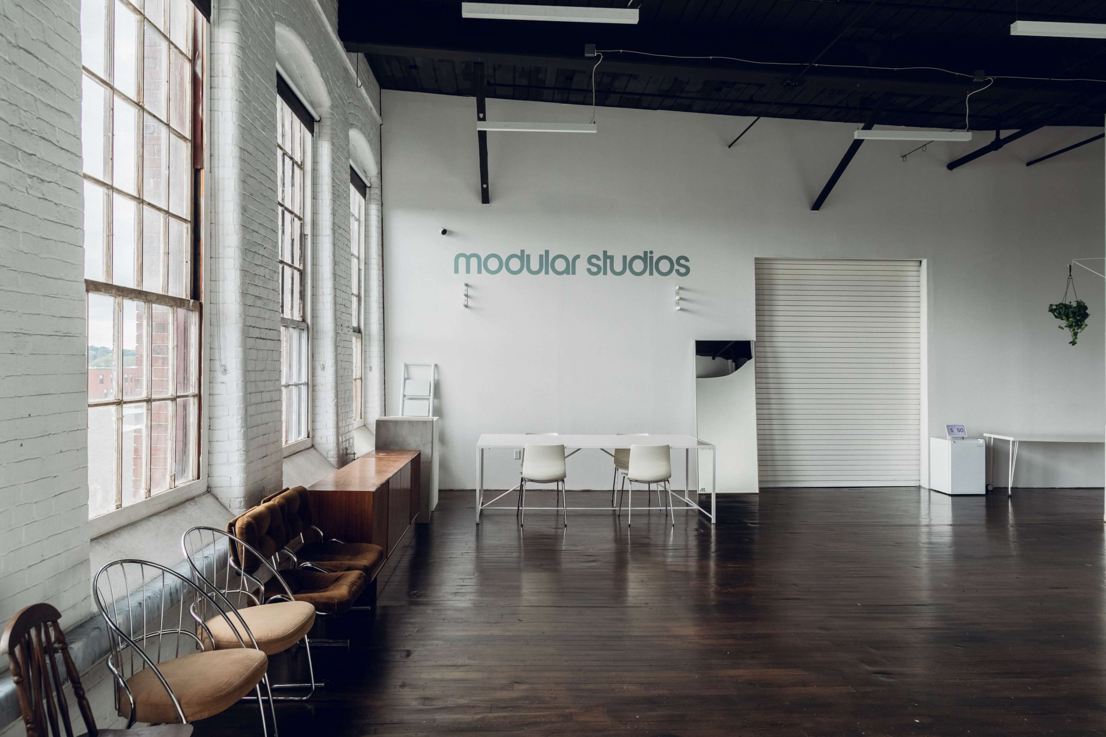 The image shows a spacious, brightly lit studio with large windows, a white wall featuring the text "modular studios," and a minimalist furniture arrangement including several chairs and a table.