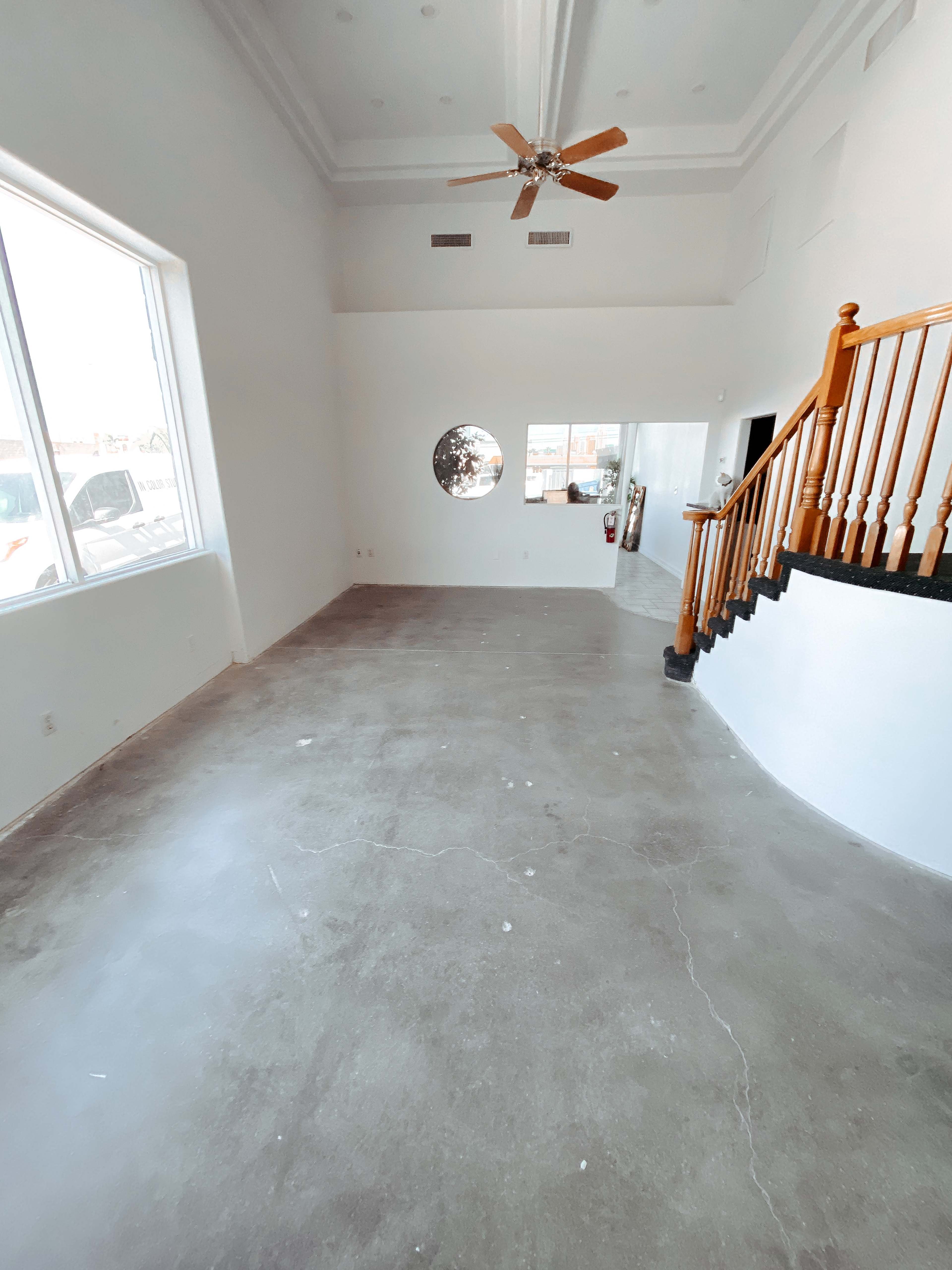 A spacious, empty room with polished concrete flooring, large windows, and a staircase with wooden railings.