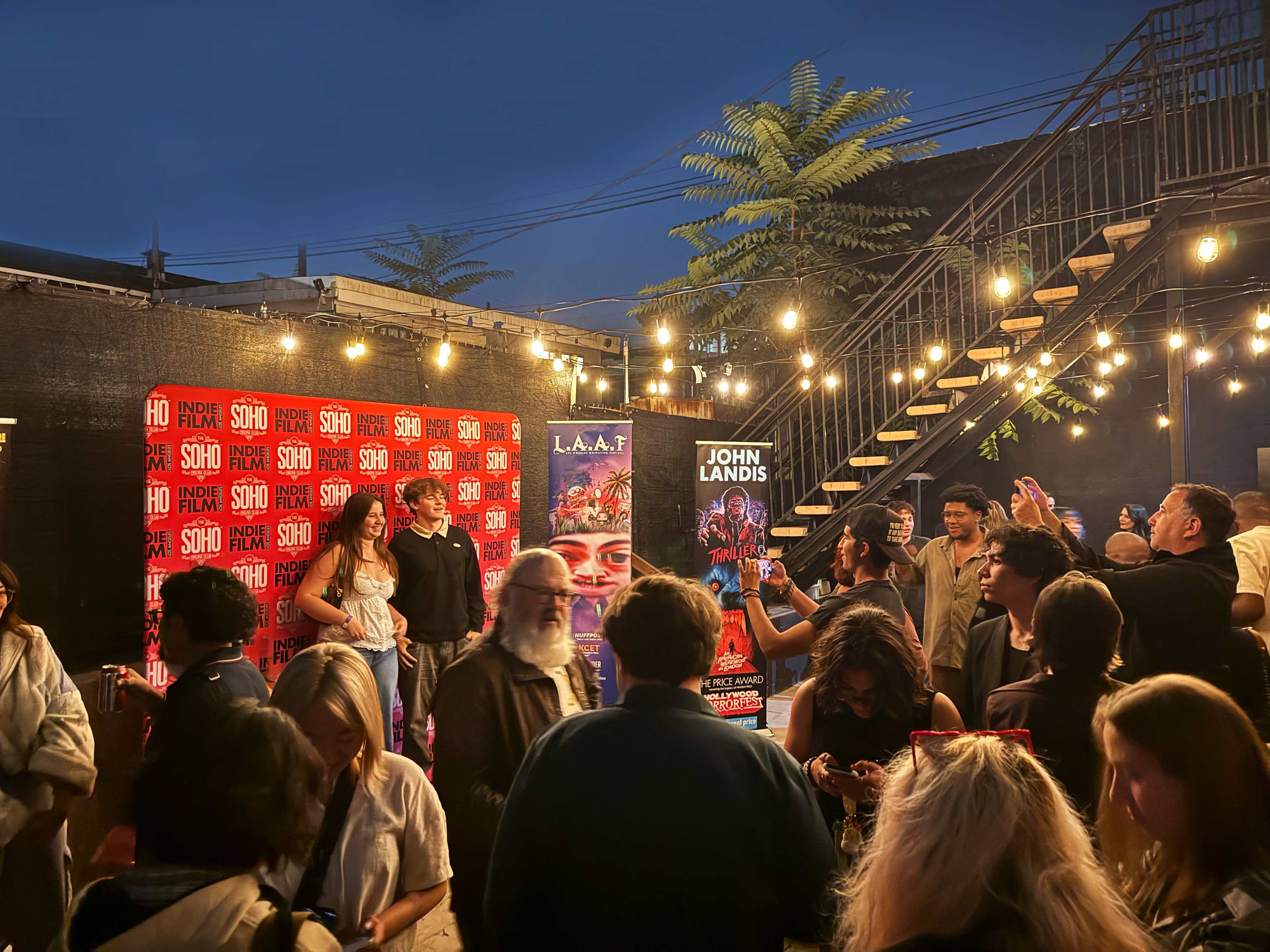 A crowded outdoor event is taking place at dusk, featuring a red backdrop for photo opportunities and string lights illuminating the area.