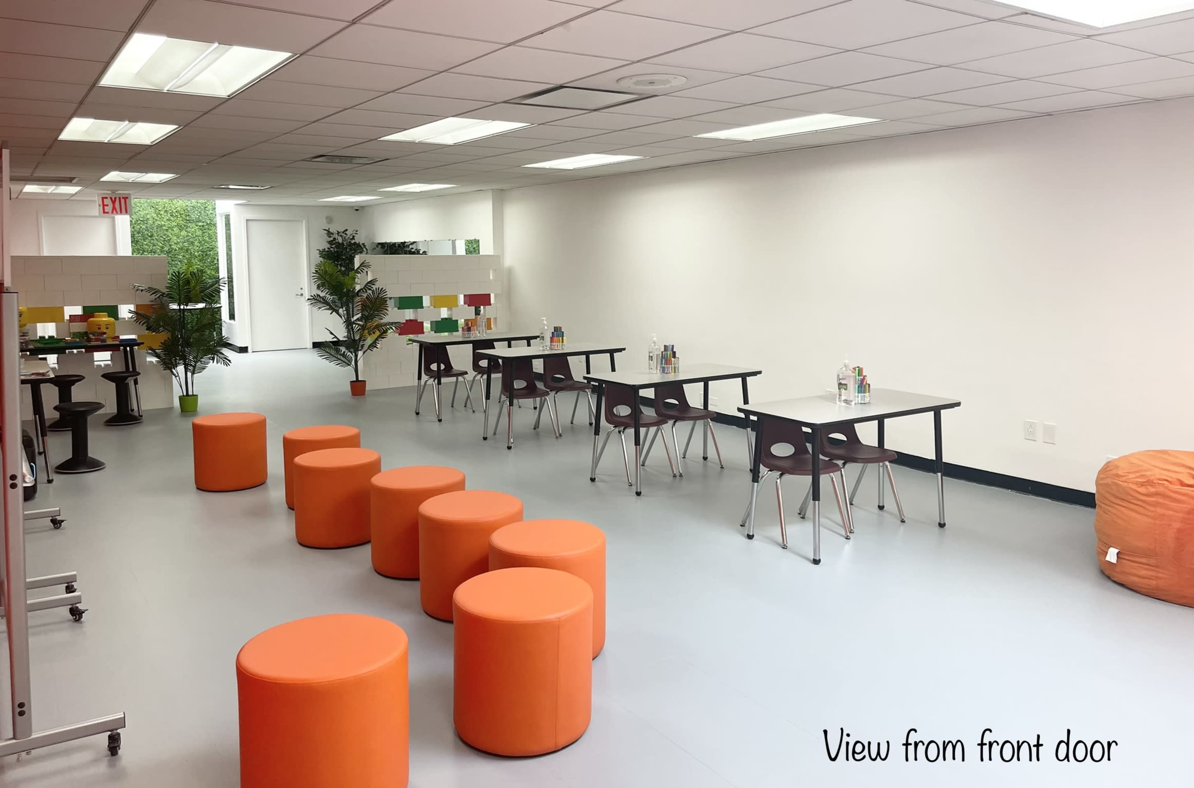 A spacious, well-lit room with several tables, orange stools, and potted plants arranged neatly along the walls.