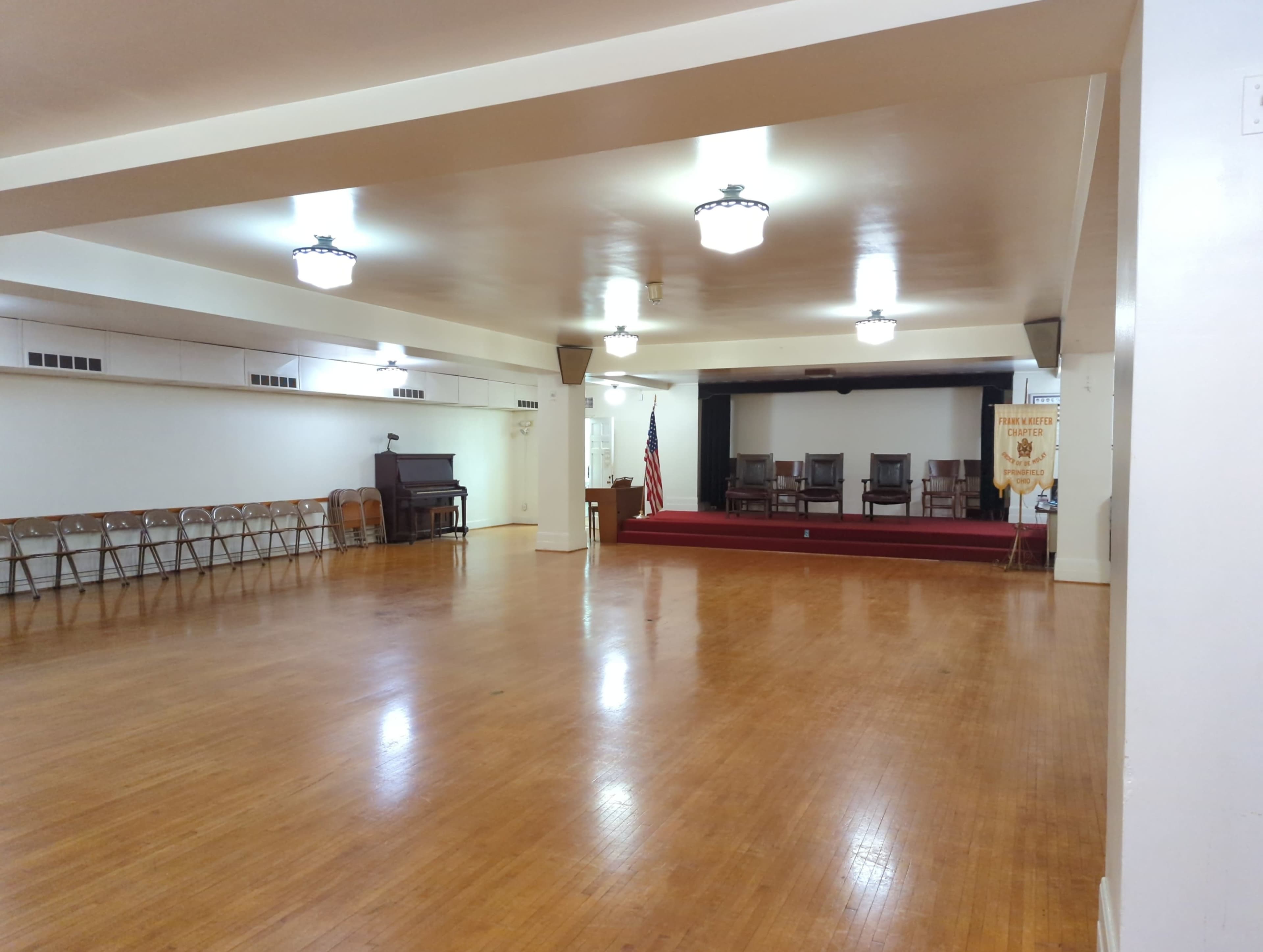 A spacious hall with wooden floors, a set of empty chairs lined against one wall, a podium at the front, an American flag, and a small stage with three chairs on a raised platform.