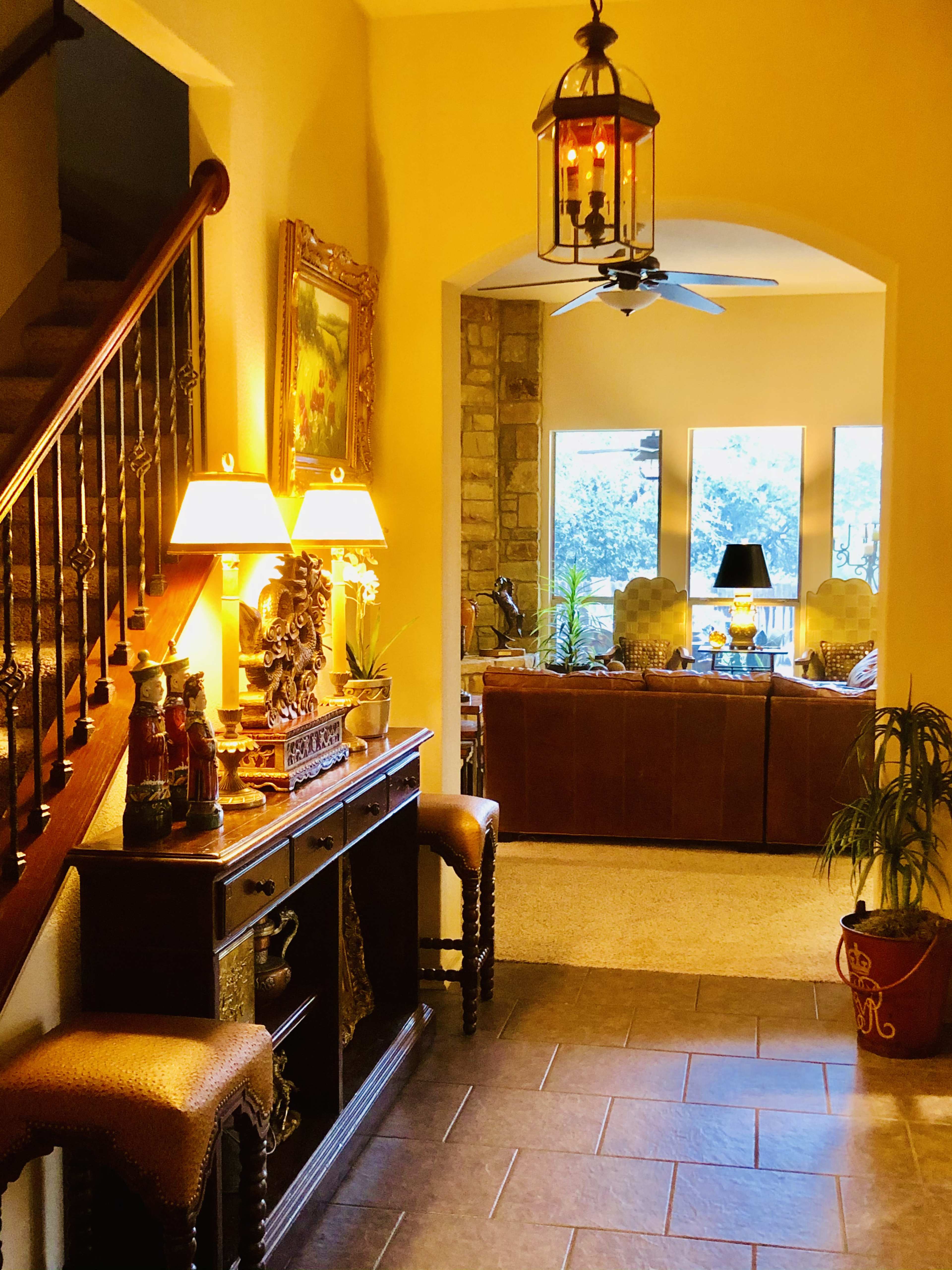 A hallway leads to a living room, featuring a staircase, decorative lighting, and a console table with floral arrangements.