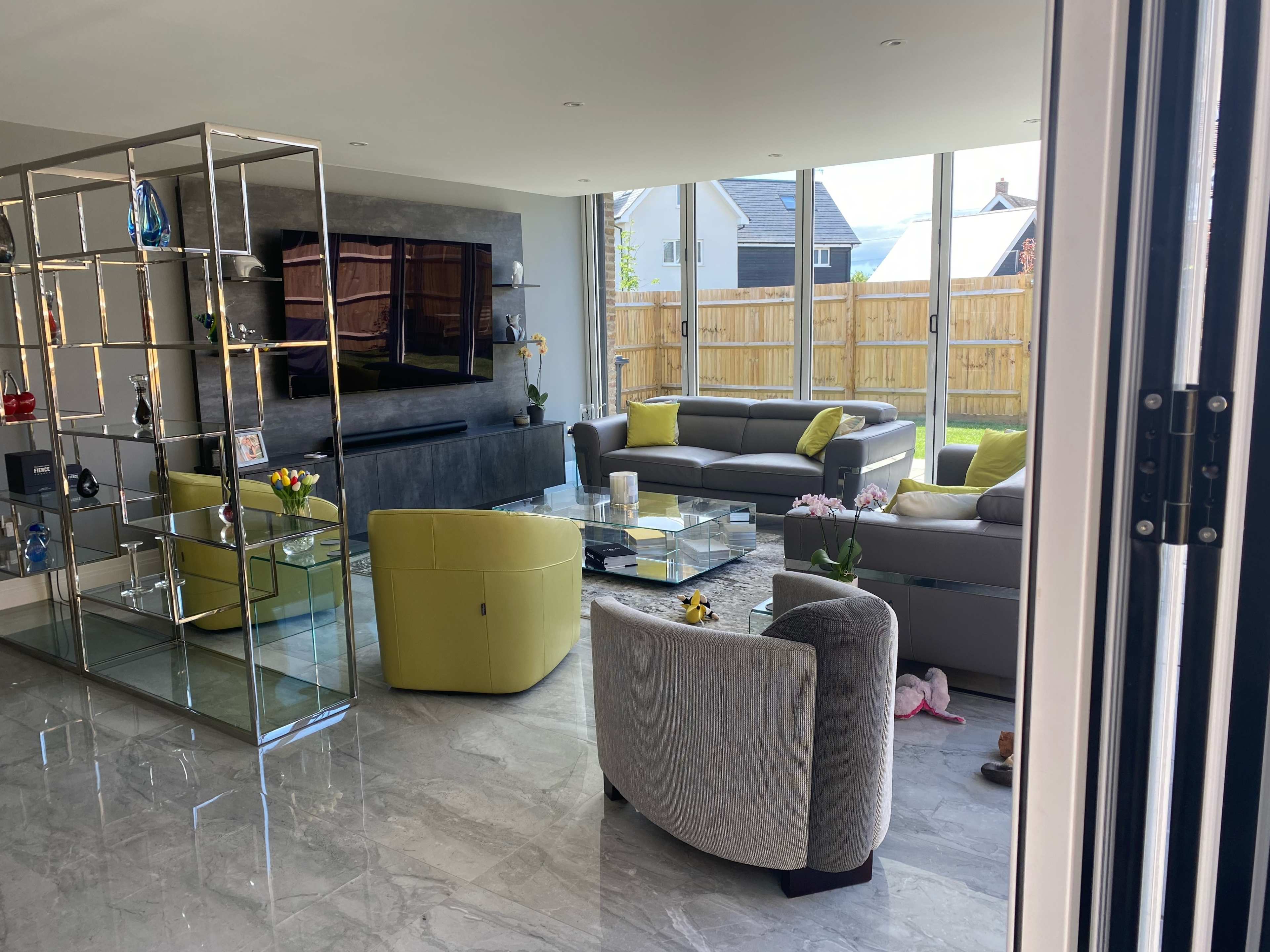 A spacious living room features modern furniture, a television mounted on the wall, and large windows overlooking a fenced yard.