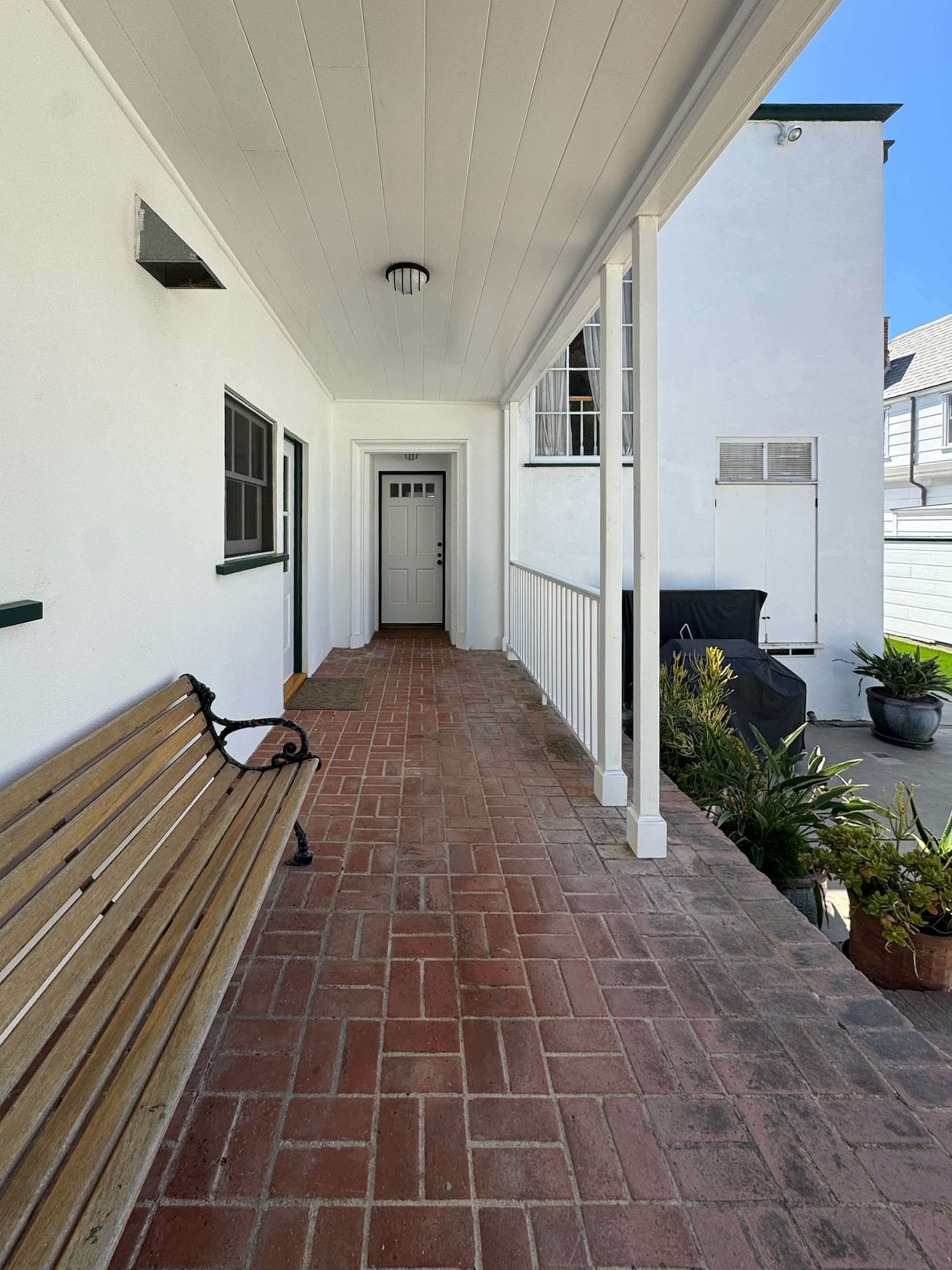 The image shows a brick pathway leading to a white door, with a wooden bench on one side and potted plants along the edges.
