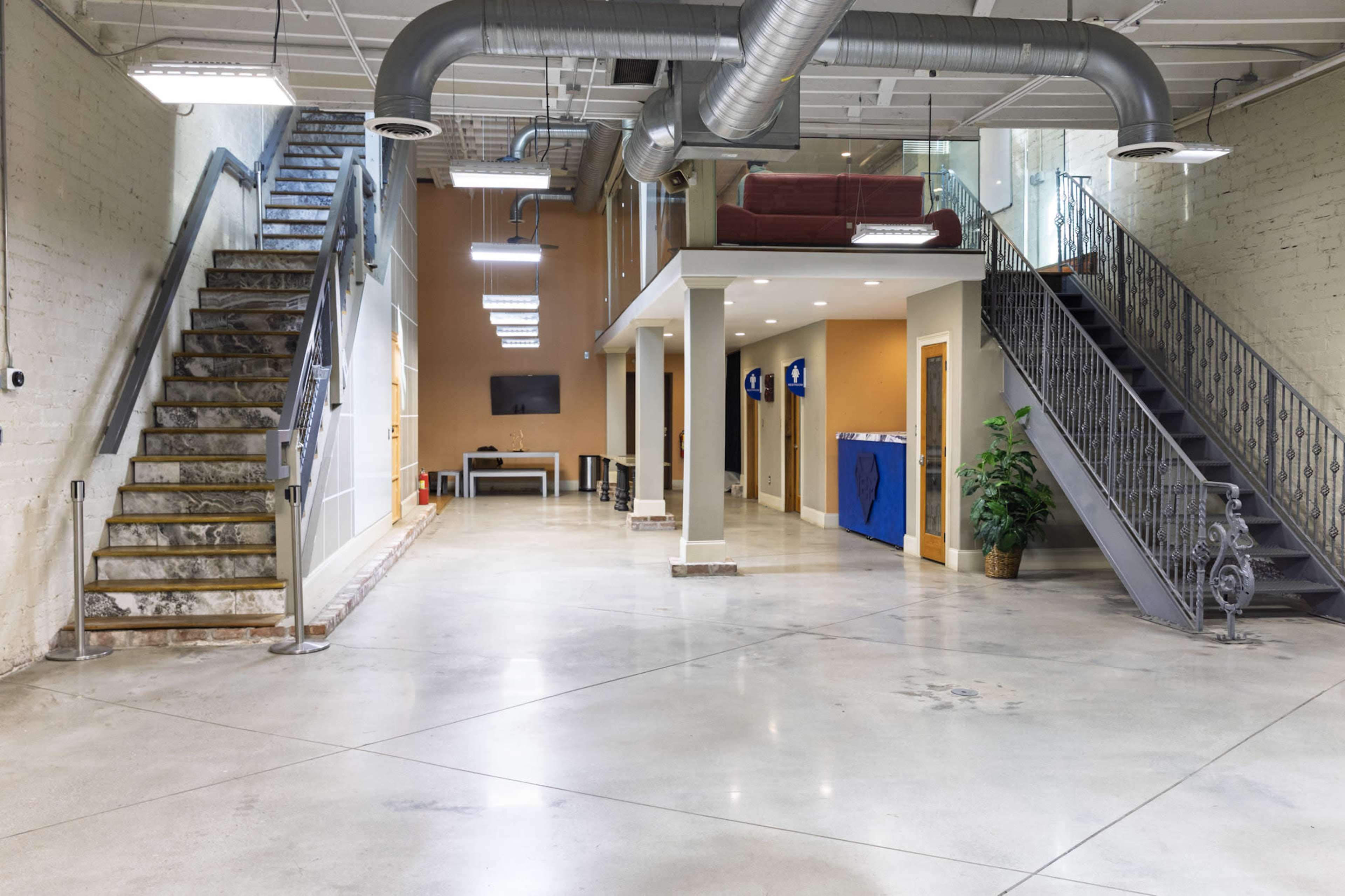 The interior of a spacious building features two staircases leading to an upper level, with a lounge area and modern decor visible in the background.