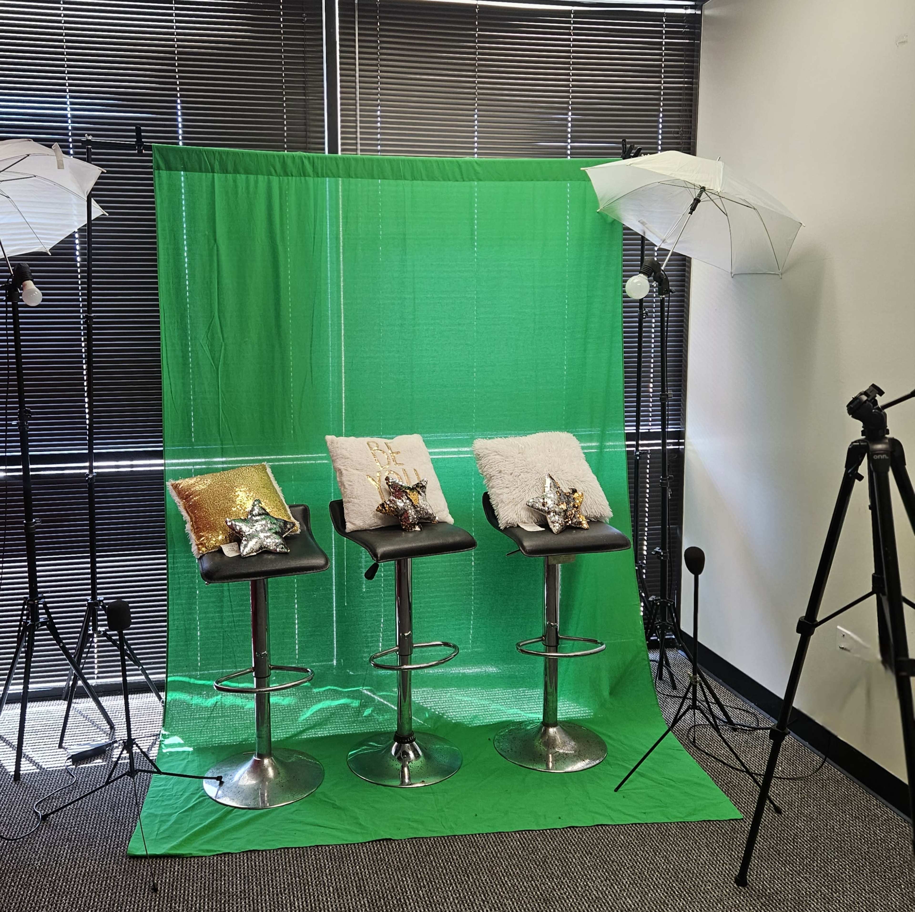 The image shows three black bar stools with decorative pillows in front of a bright green screen, with lighting equipment and a tripod positioned nearby.