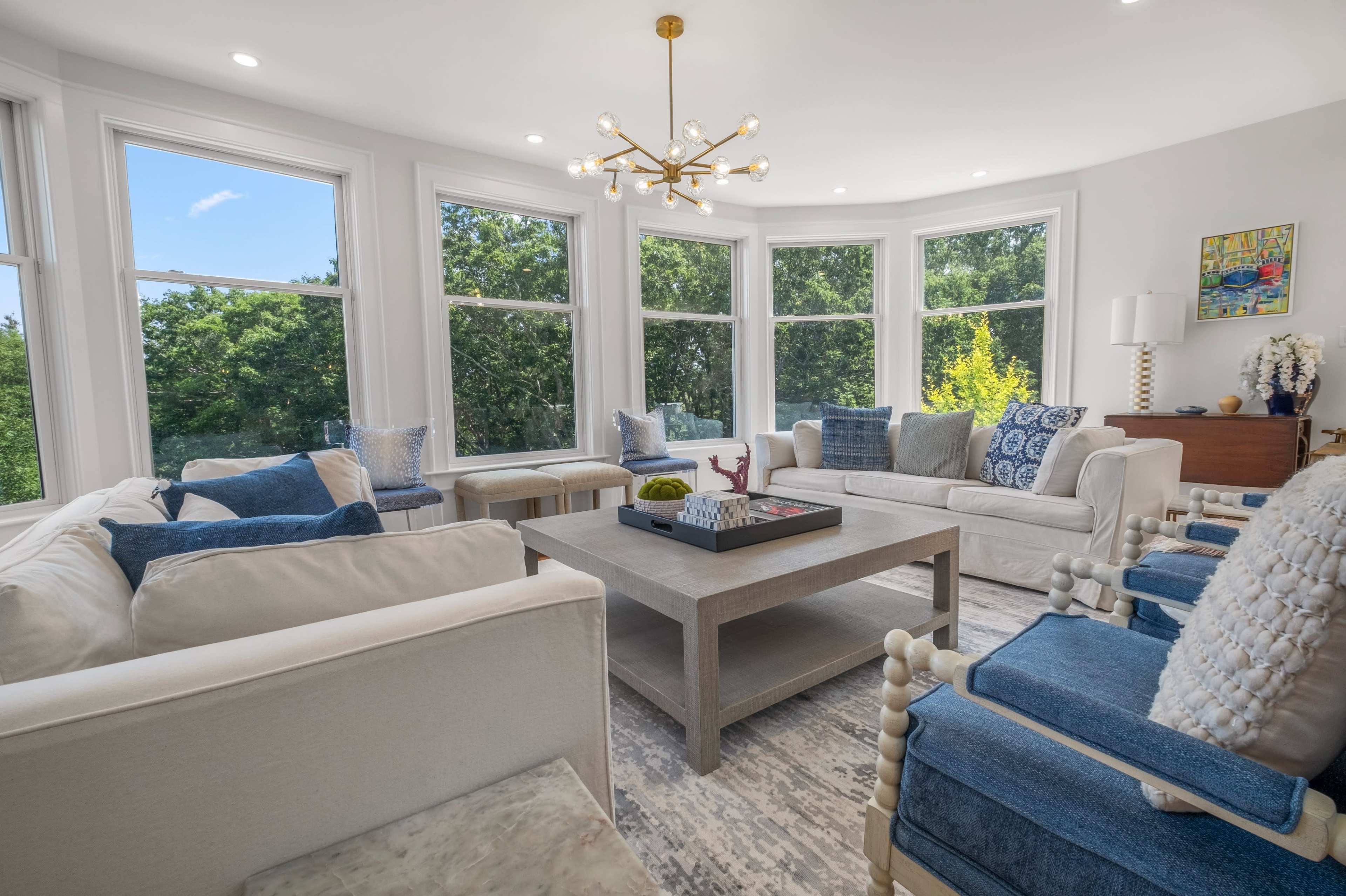 A bright, modern living room with large windows, white sofas, and blue accent chairs arranged around a central coffee table.