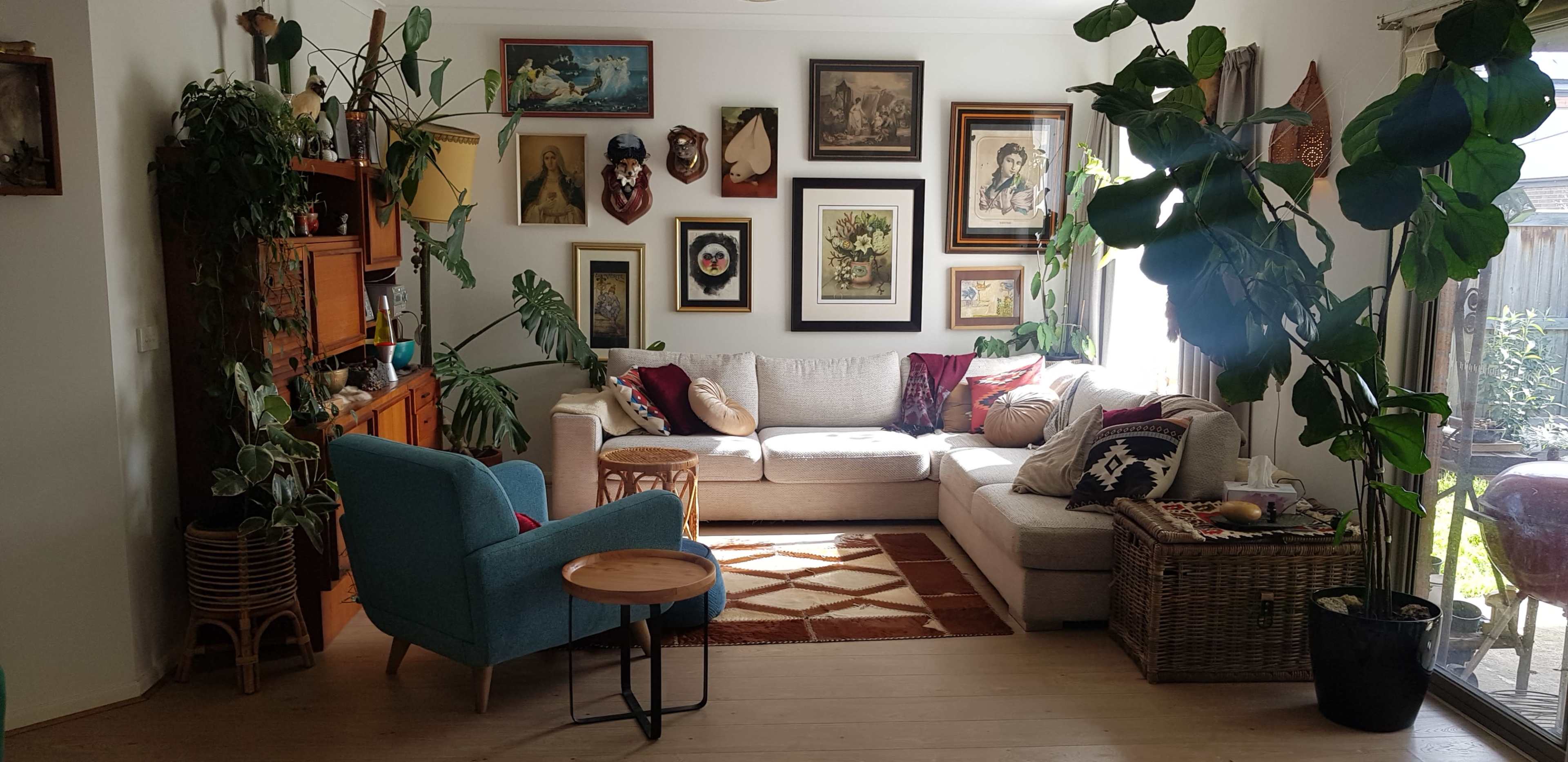 A cozy living room featuring a sectional sofa, a blue accent chair, an array of framed art on the walls, and several indoor plants.