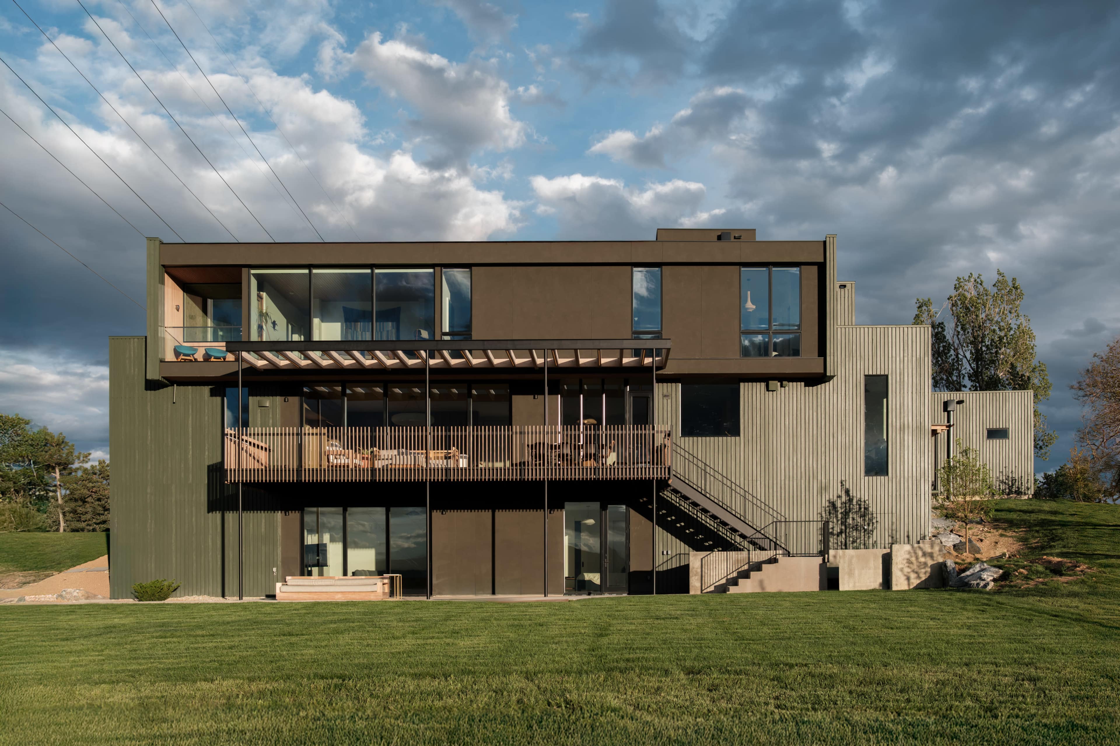 The image shows a modern, two-story house with a combination of wood and metal siding, large windows, and a balcony overlooking a grassy lawn.