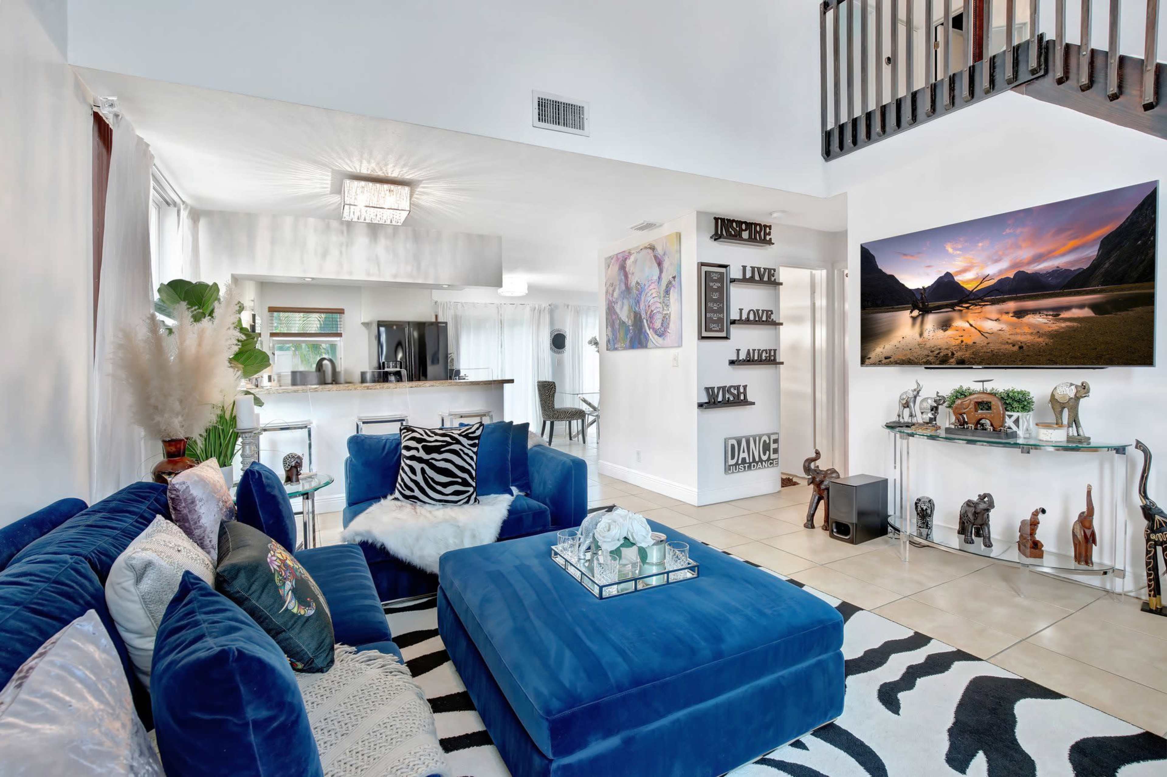 The living area features a blue velvet sectional sofa, a patterned rug, and a large flat-screen TV mounted on the wall, with kitchen and dining spaces visible in the background.
