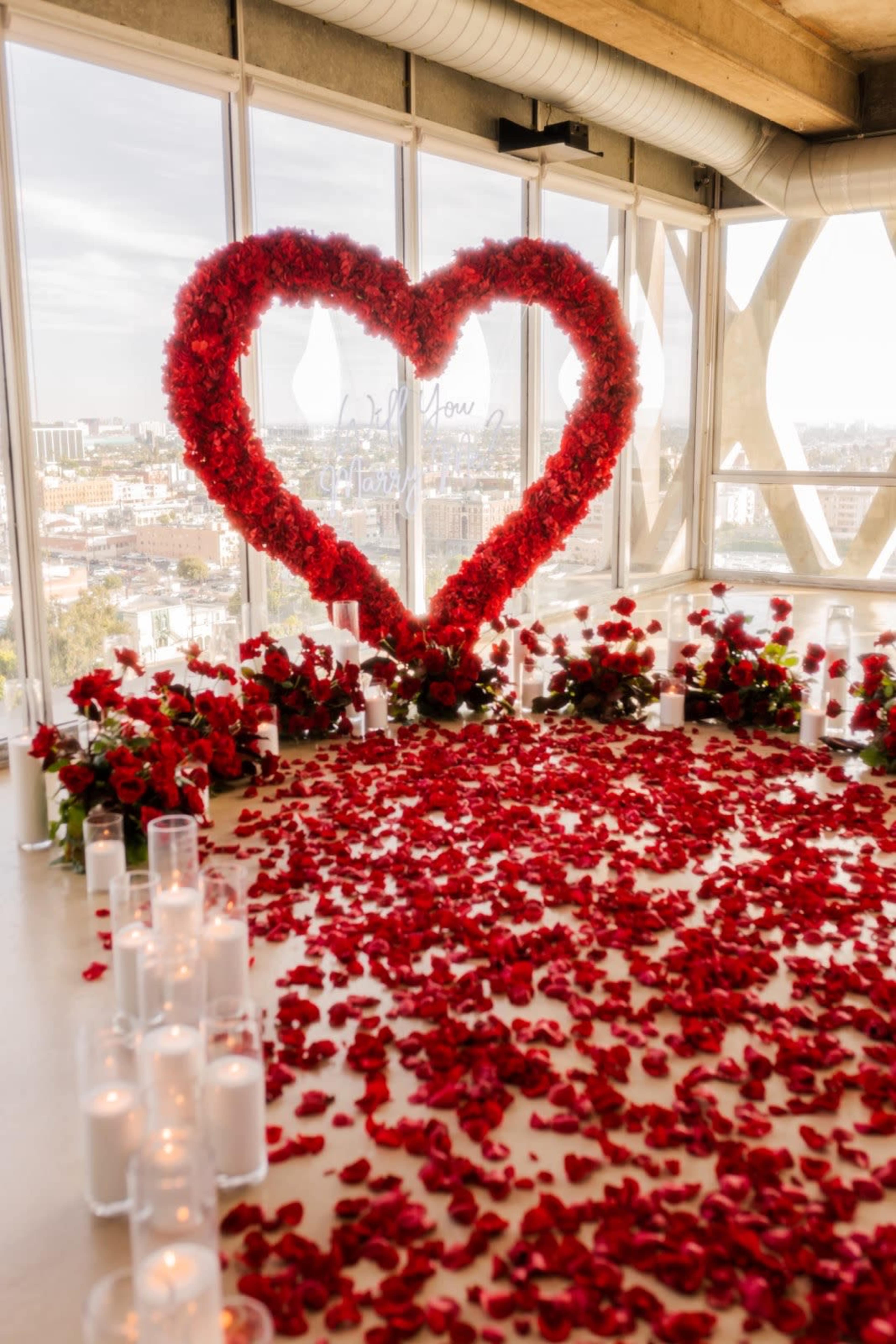 The scene features a large heart-shaped arrangement made of red flowers, surrounded by a carpet of rose petals and illuminated by candles, set against a backdrop of a city skyline through large windows.