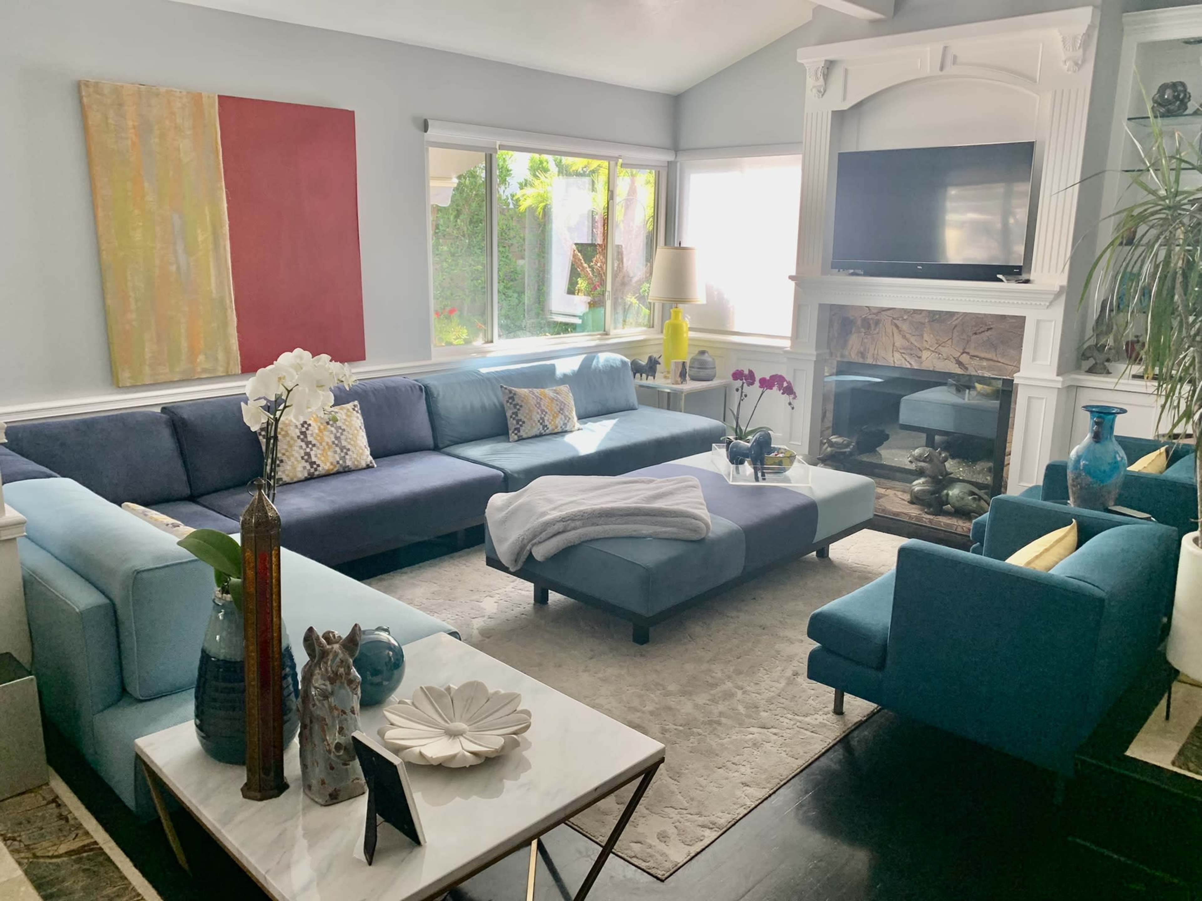 The living room features a large blue sectional sofa, a white coffee table, and a stone fireplace, with windows providing natural light and plants visible outside.