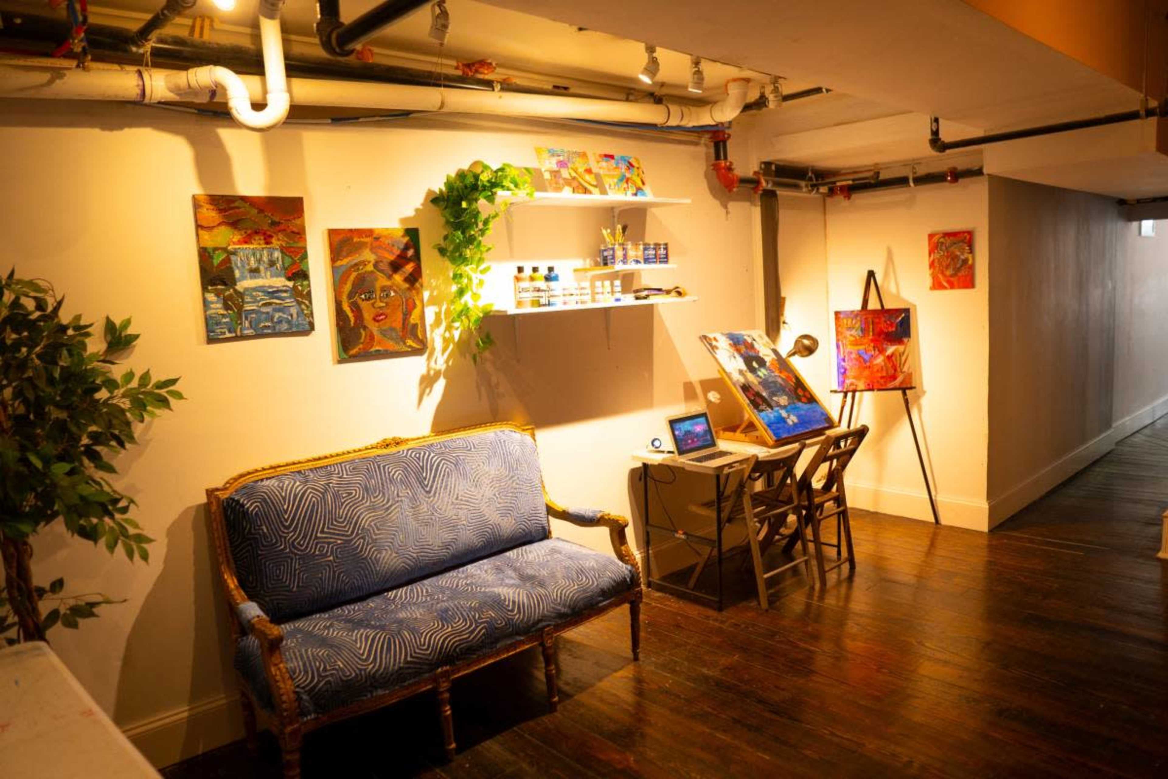 A framed art space featuring a blue sofa, a desk with an easel, colorful paintings on the walls, and shelves holding art supplies.