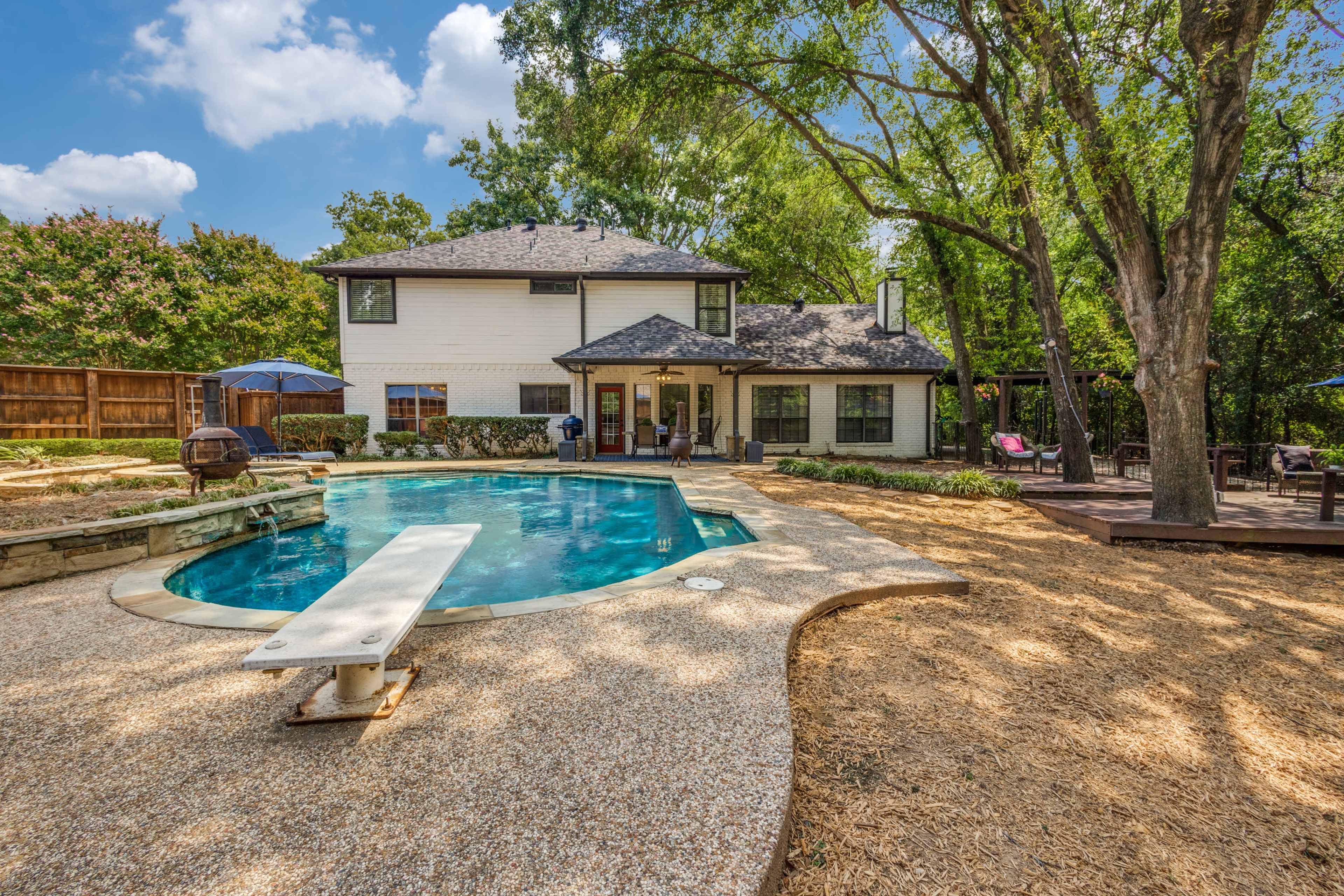 The image shows a backyard featuring a swimming pool with a diving board, a two-story house, and shaded areas with trees and landscaping.